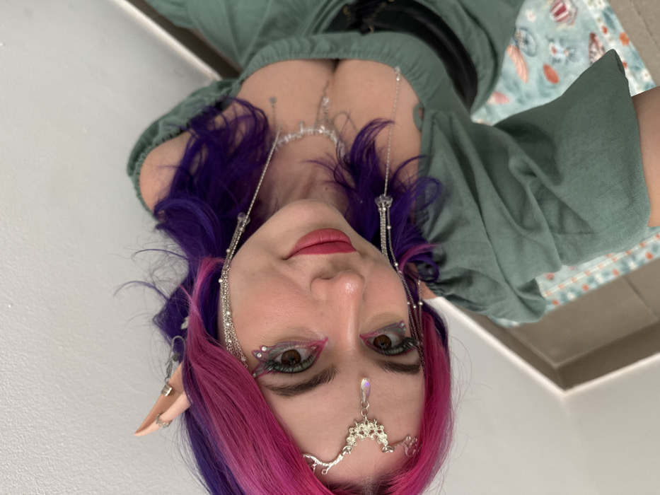 A woman with pink and purple hair, pointed elf ears, and decorative jewelry poses for the camera. She wears a light green top with a dark belt. Her makeup includes winged eyeliner and a gem on her forehead. She has a fair complexion and red lipstick. A necklace adorns her chest. The background is a mix of a wall and a patterned surface.