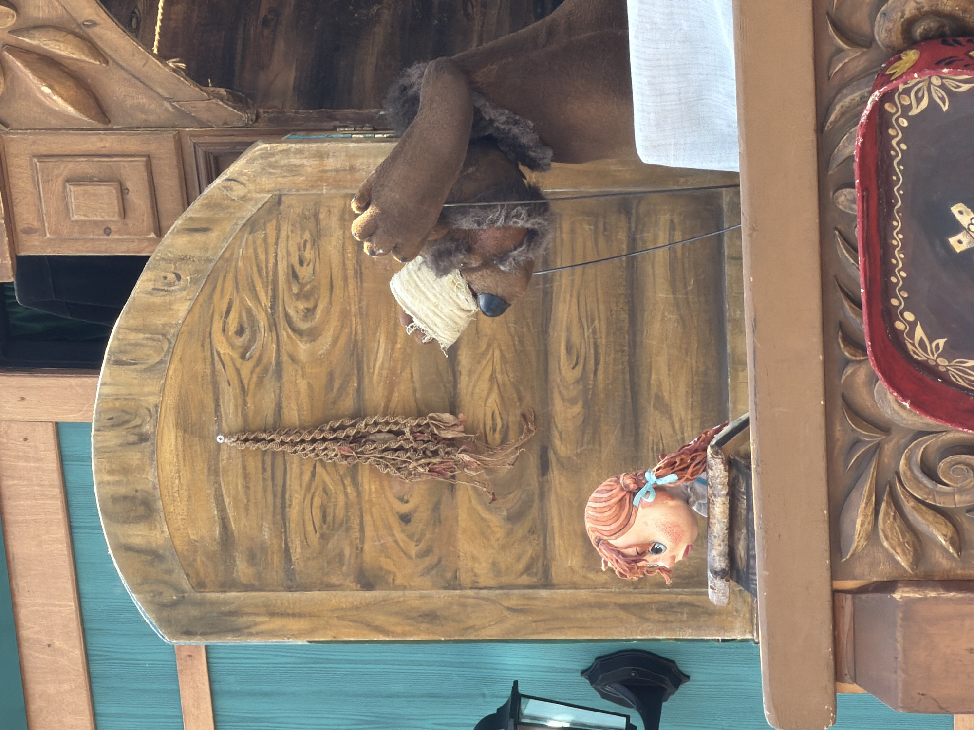 The image presents an animatronic or puppet display featuring two characters within a rustic, wooden-themed setting. On the right, a large, brown, furry animal figure, possibly a bear, is positioned with a white bandage around its head. Its right arm is raised, covering its eyes or forehead, conveying an impression of distress, shyness, or deep thought. A thin, dark control rod extends downwards from its head. To the left and slightly below, a smaller, human-like doll with reddish-brown hair styled in pigtails, secured with light blue bows, looks towards the right. Her wide blue eyes and slightly open mouth suggest an expression of surprise or concern. She wears a light-colored garment.
The background consists of a painted wooden backdrop designed to resemble horizontal planks, topped by a curved arch-like structure. A small, dried bundle of reddish-brown stringy material hangs from a hook on this backdrop, situated above and to the left of the doll. To the far left, a teal-colored wall with horizontal paneling is visible, partially obscured by a black wall-mounted lantern. To the far right, behind the animal figure, a dark fabric, likely a curtain, is visible through an opening, alongside intricate carved wooden architectural details. The characters are situated on a multi-layered wooden platform. The immediate foreground reveals a rich, carved wooden base, with a red tray or plate decorated with gold patterns (including swirls, leaves, and a central cross-like design) partially visible on the bottom right.
The image provides no identifiable visual cues to confirm its location in Khimki, Russia. The scene appears to be a staged display, likely from an amusement park, museum, or performance venue.