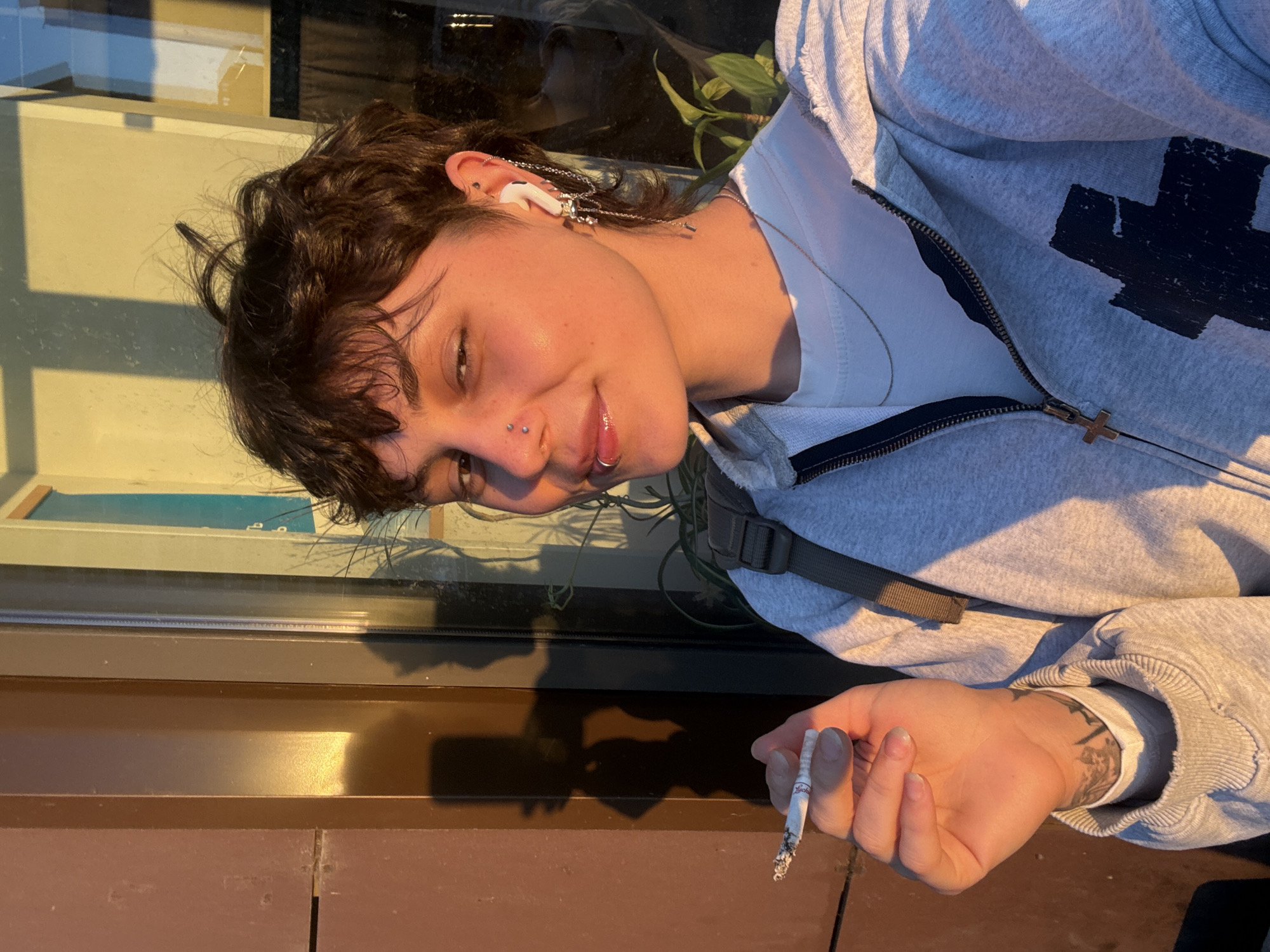 A person with short, dark, curly hair, a nose ring, and a lip ring, is taking a selfie outdoors at golden hour. They are holding a lit cigarette in their left hand and are wearing a light grey zip-up hoodie over a white t-shirt, a silver necklace, and a dangling earring in their right ear. The background shows a building with a brown facade and a large window, reflecting sunlight. Plants are visible behind the person through the window. The image was taken in Saint Petersburg, Russia.