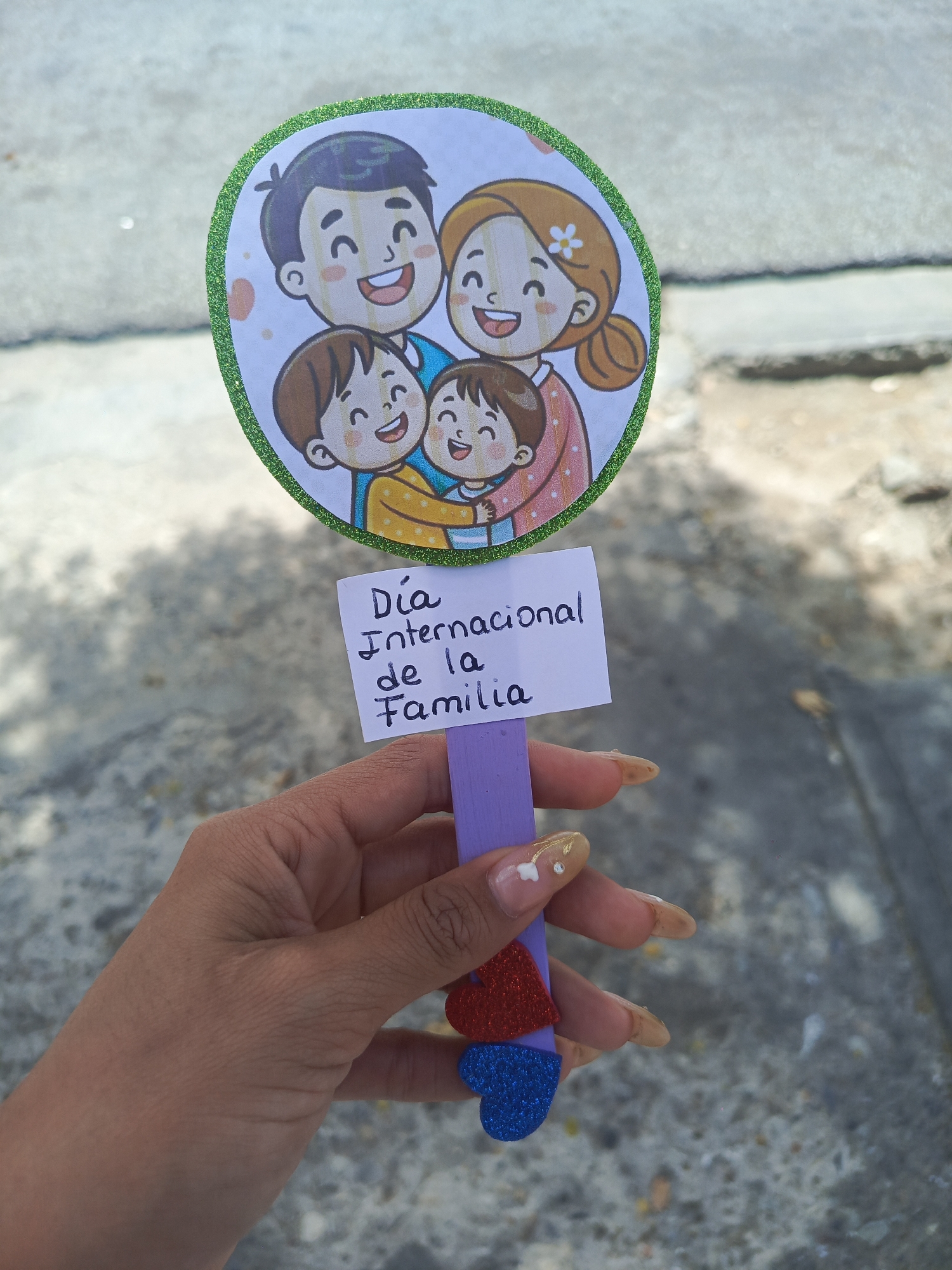 A hand holding a craft project is in the foreground. The background is of a city street or sidewalk.
The craft project consists of a drawing of a cartoon family with two parents and two children, encased in a green glittery border. Below the cartoon family is a small rectangular piece of paper with the inscription "Día Internacional de la Familia," attached to a purple popsicle stick. Two glittery hearts are attached at the bottom of the popsicle stick, one red and one blue.
The hand holding the craft project has long fingernails that are painted with a nude color. One fingernail has a small white floral design. The street or sidewalk in the background is a neutral gray color.
No specific information in this image indicates that the location is in the city of Juan Griego, Venezuela.