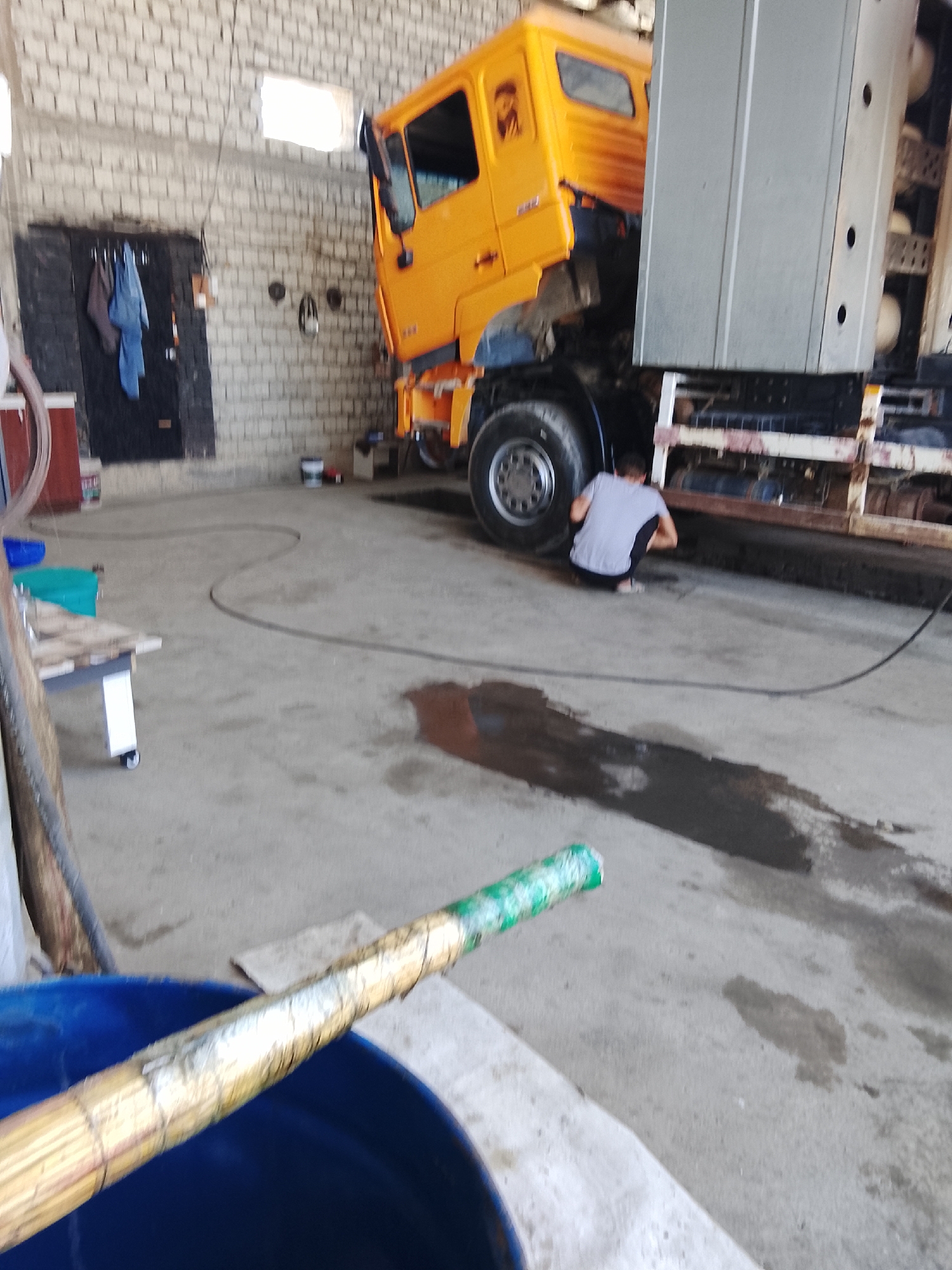 In Sabir Rakhimov, Uzbekistan, an orange truck is parked in a garage with a concrete floor and brick walls. A person in a grey t-shirt and black pants is squatting near the rear wheel of the truck. A large blue container with a wooden tool is in the foreground. Various objects, including buckets, boxes, and tools, are scattered around the garage. The scene appears to be a workshop or maintenance area.