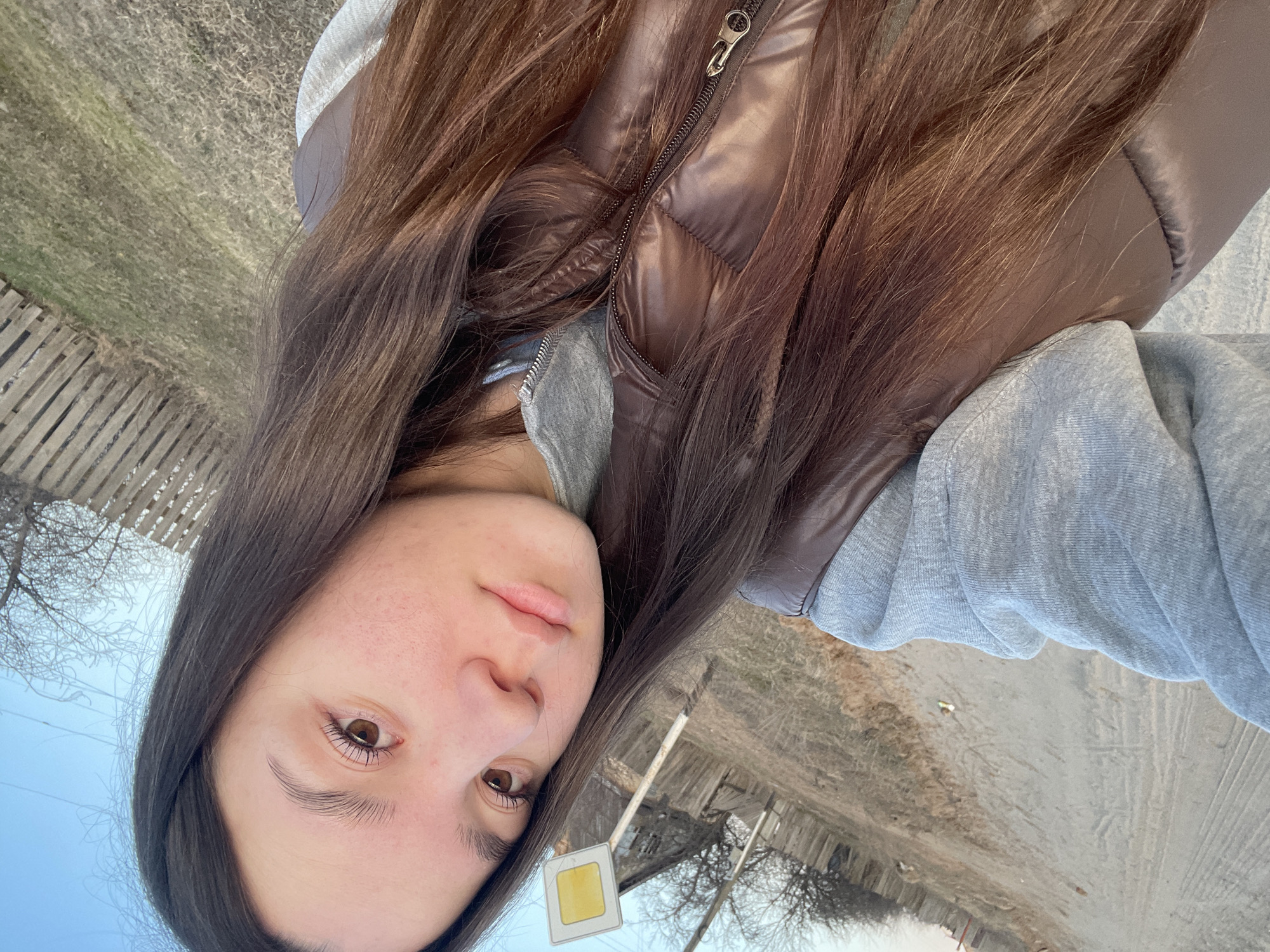 This outdoor photograph features a young woman looking directly at the camera. She has long, straight dark brown hair that falls over her shoulders and chest. Her expression is neutral, with a slight pout to her lips, and her dark brown eyes are open. Her skin appears natural with some minor visible blemishes.
She is wearing a glossy, dark brown or possibly reddish-brown puffer vest unzipped over a light grey hooded sweatshirt.
The background reveals a rural or semi-rural setting. On the left side, there's a rustic wooden fence. Beyond it, the ground is covered with dry, light-colored grass and patches of green, indicative of a field or uncultivated land. To the right, a dirt road or path with visible tire tracks extends into the distance. Bare tree branches are silhouetted against a clear, light blue sky, suggesting the season is late autumn, winter, or early spring.
A square yellow road sign with a white border is visible in the lower-central part of the image, mounted on a wooden post. Utility wires can be seen against the sky in the upper left.
The primary activity is the woman posing for a photograph, likely a selfie. The lighting is soft and natural, suggesting daytime, possibly late afternoon. No discernible text is visible on the sign or elsewhere in the image. The setting is consistent with the provided location context of Ольховка, Russia.