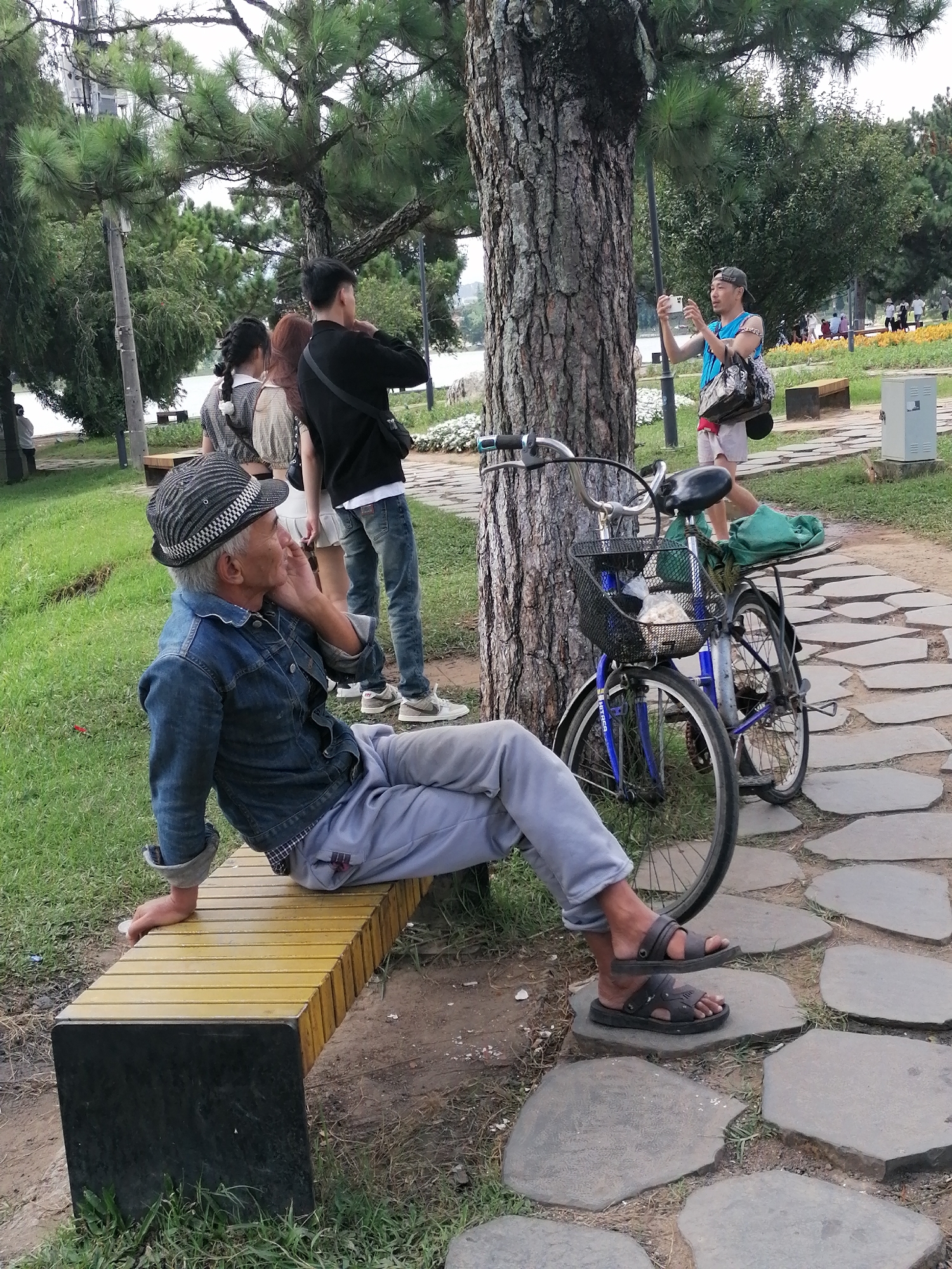 In Da Lat, Vietnam, a man in a denim jacket, grey pants, and a patterned hat sits on a yellow and black bench, facing left, with his hand on his chin. His sandals are on the ground. Behind him, a blue bicycle with a basket and a green bag rests by a tree trunk. Further back, a man in a blue shirt and shorts holds a phone to take a picture. Two women and a man in a black shirt stand in the background near a paved path and other parkgoers. The scene is set in a park with green grass, trees, and a stone pathway.