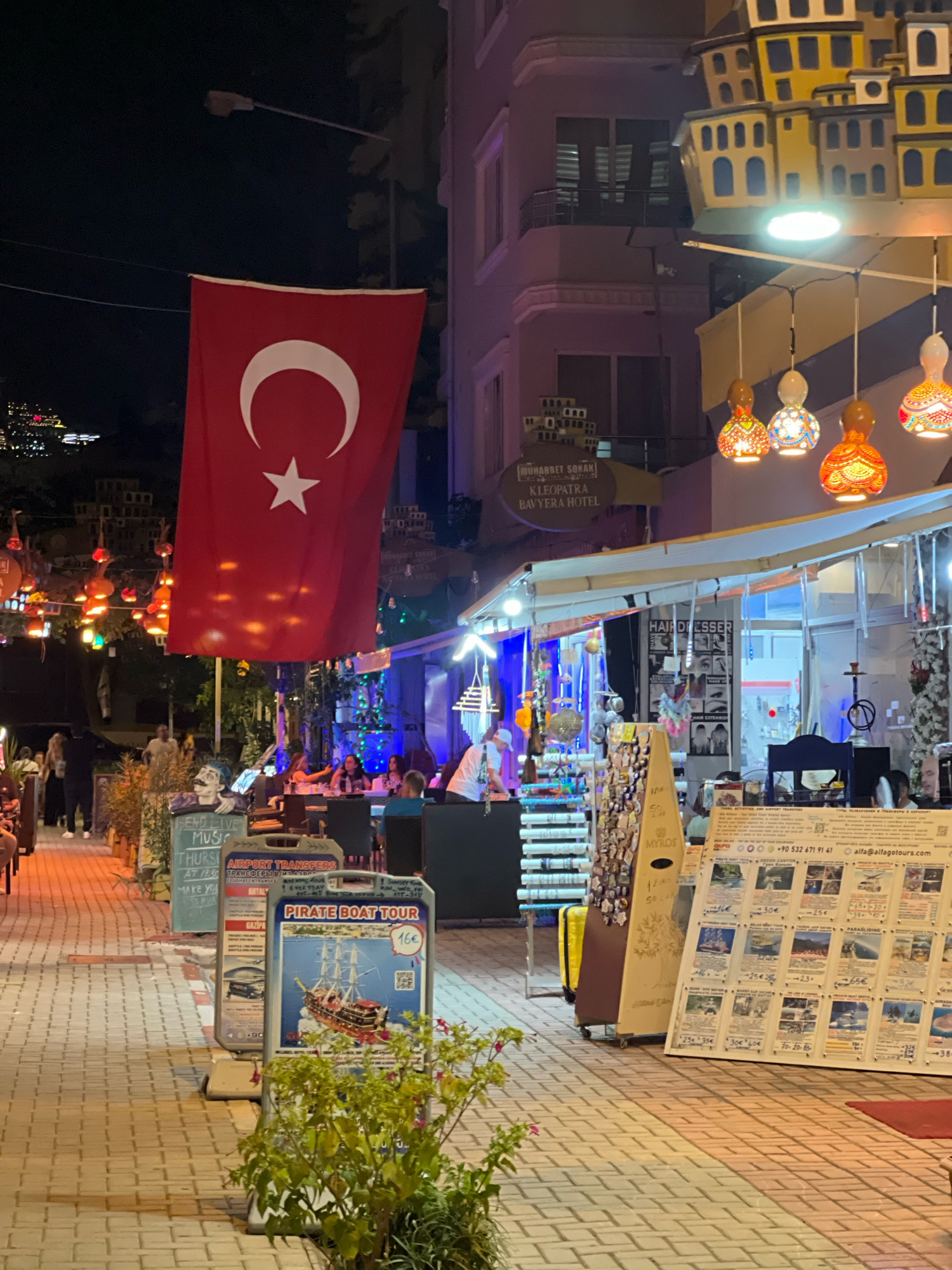 The image depicts a nighttime scene on a pedestrian walkway in Alanya, Turkey, likely a popular tourist area. A large Turkish flag hangs prominently in the center, partially obscuring a building adorned with decorative, yellow, multi-story structures resembling houses. Below the building, several shops with awnings are visible, displaying goods and illuminated signs. Colorful, gourd-shaped lanterns hang from wires, casting a warm glow.
On the left side of the walkway, a street lamp is visible above the flag. Further down the walkway, people are seated at tables outside what appears to be a cafe or restaurant. Signs are displayed along the walkway, including one advertising "PIRATE BOAT TOUR" with prices in Euros, and another listing "AIRPORT TRANSFERS." A rack of keychains and souvenirs is also visible. The walkway is paved with light-colored bricks. A small bush with green leaves and pink flowers grows in the foreground. The overall atmosphere suggests a lively evening in a tourist district.