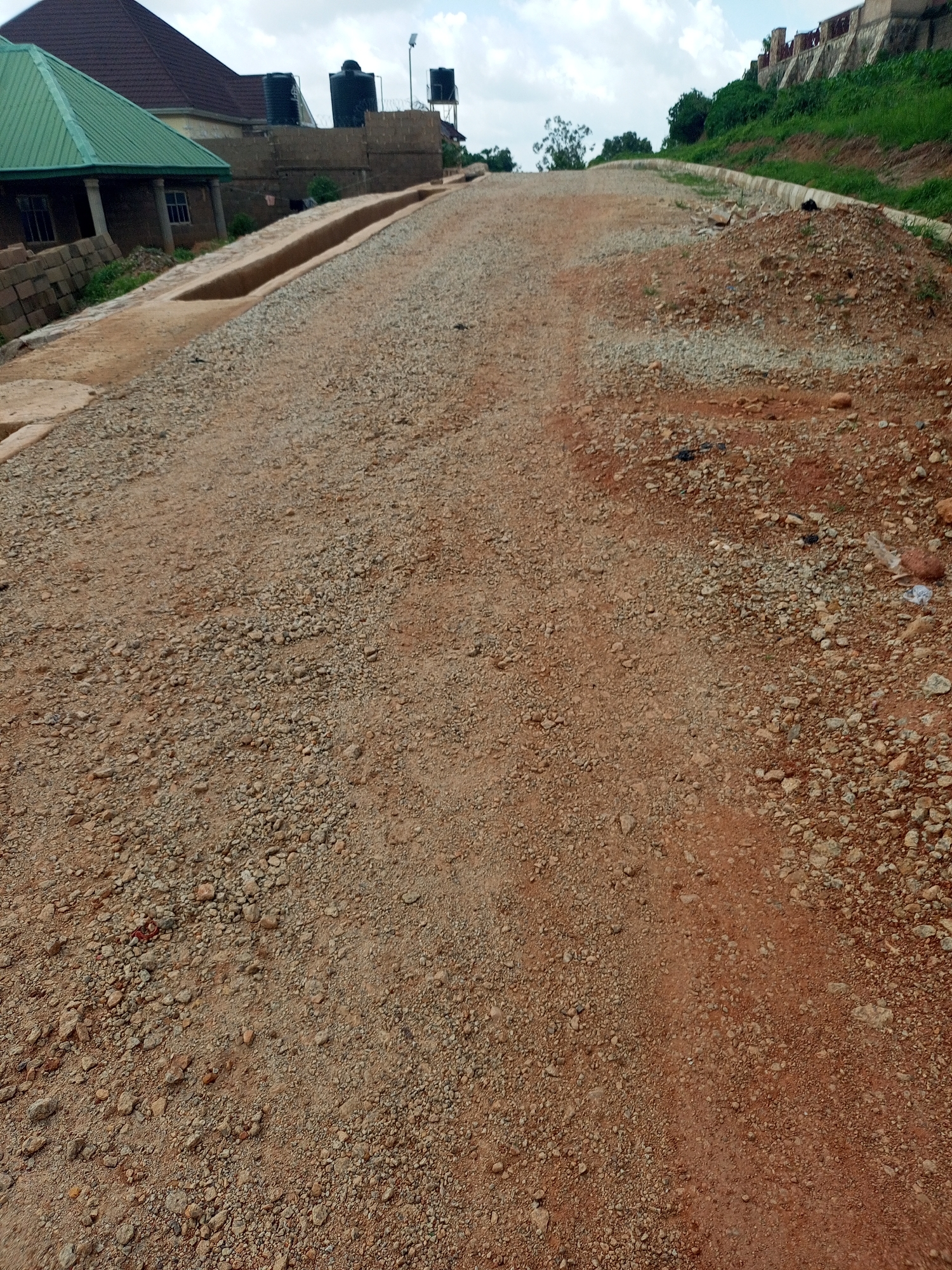 A dirt road curves upwards through a construction site in Jos, Nigeria. The road is made of reddish-brown soil mixed with gravel. To the left, a building with a green roof and several windows is partially visible, alongside stacks of bricks and a concrete drainage channel. In the background, two large black water tanks are mounted on a raised platform, and a solar-powered street light is visible. The road is flanked by slopes with green vegetation and a concrete curb on the right side. The sky is cloudy, with patches of blue. The scene depicts ongoing infrastructure development in an urban or semi-urban area.