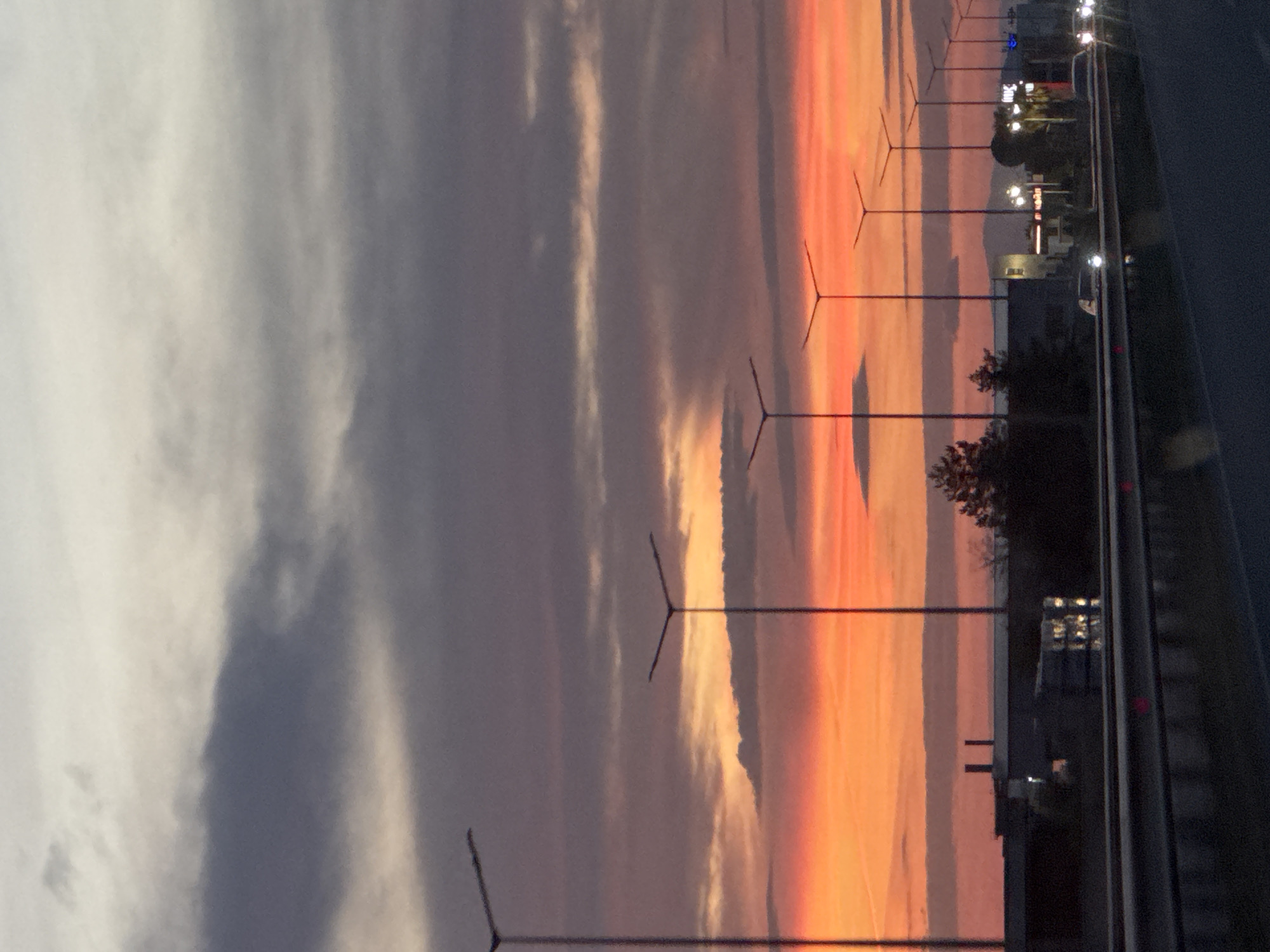 The image shows a sunset scene in Asenovgrad, Bulgaria. The sky is filled with clouds, with shades of gray and white on the left side transitioning to orange and red on the right side where the sun is setting. A row of street lights is visible along the right side of the image, bordering a highway. Buildings and trees can be seen in the background, near the streetlights. The image appears to be taken from a moving vehicle.
