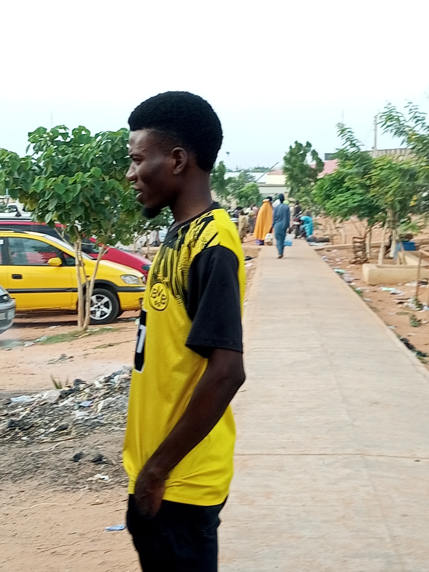 In the city of Gombe, Nigeria, a young Black man stands facing left, his profile visible. He has short, dark, curly hair and is wearing a bright yellow t-shirt with black sleeves and black trim around the neckline. The t-shirt features a black design on the front with a circular emblem. He is also wearing black pants. His left hand is tucked into his pants pocket.
Behind him, a bright yellow car with a red roof is parked on a dirt surface. To the left of the car are green trees with dense foliage. Further down a paved walkway, several people are walking away from the camera. One person is wearing a bright orange or yellow headscarf and a flowing orange garment. Another person is wearing a blue robe. More trees and some structures are visible in the background. The ground in the foreground and along the sides of the walkway is a mix of dirt and scattered debris. The sky is overcast.