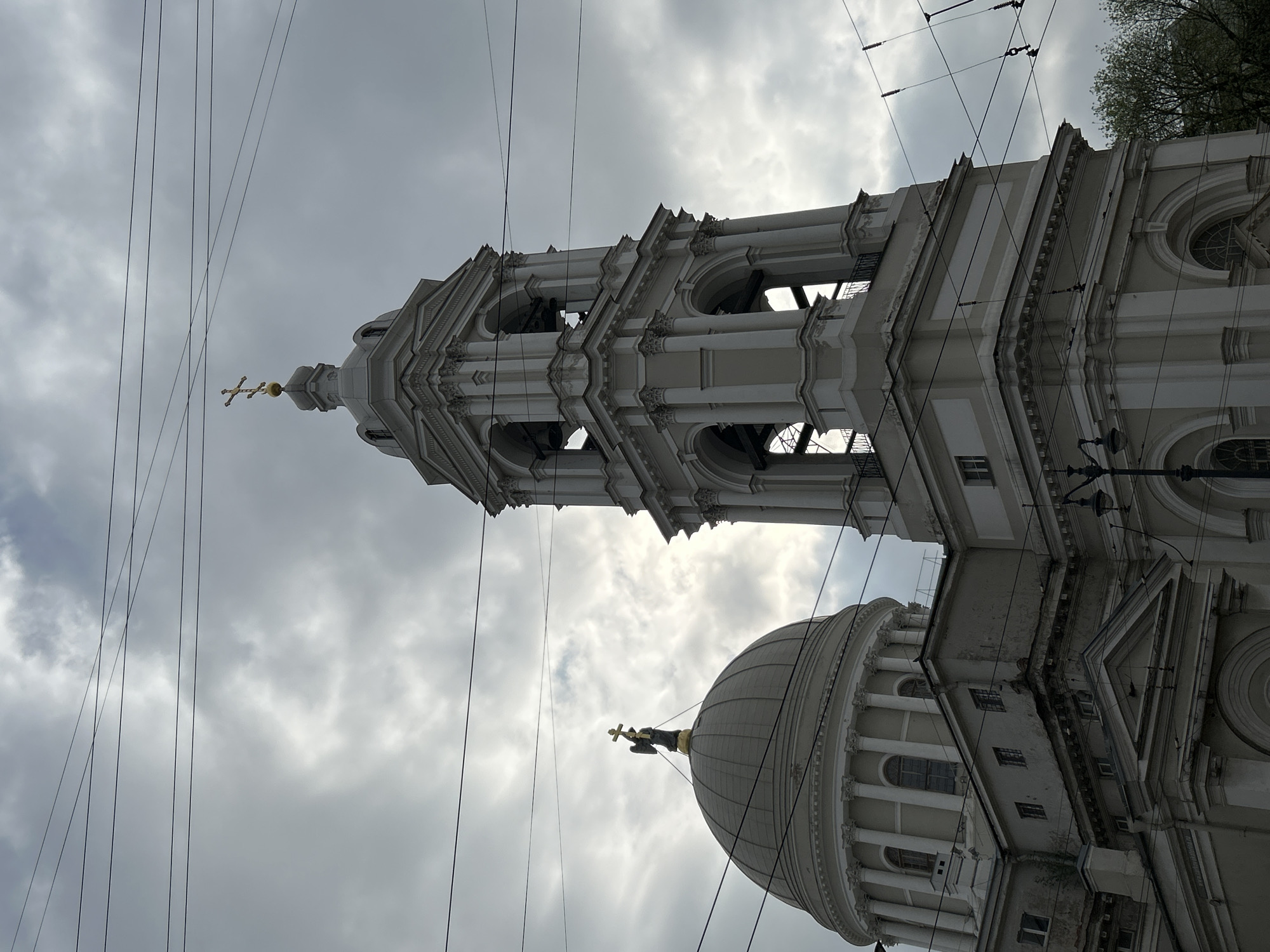 A low-angle shot shows a cathedral complex, its architectural elements contrasting against a cloudy sky intersected by power lines. The dome of the cathedral, adorned with a statue of a religious figure holding a cross, sits adjacent to a tall bell tower with open arches and decorative details, both rendered in light colors. The location is Saint Petersburg, Russia.