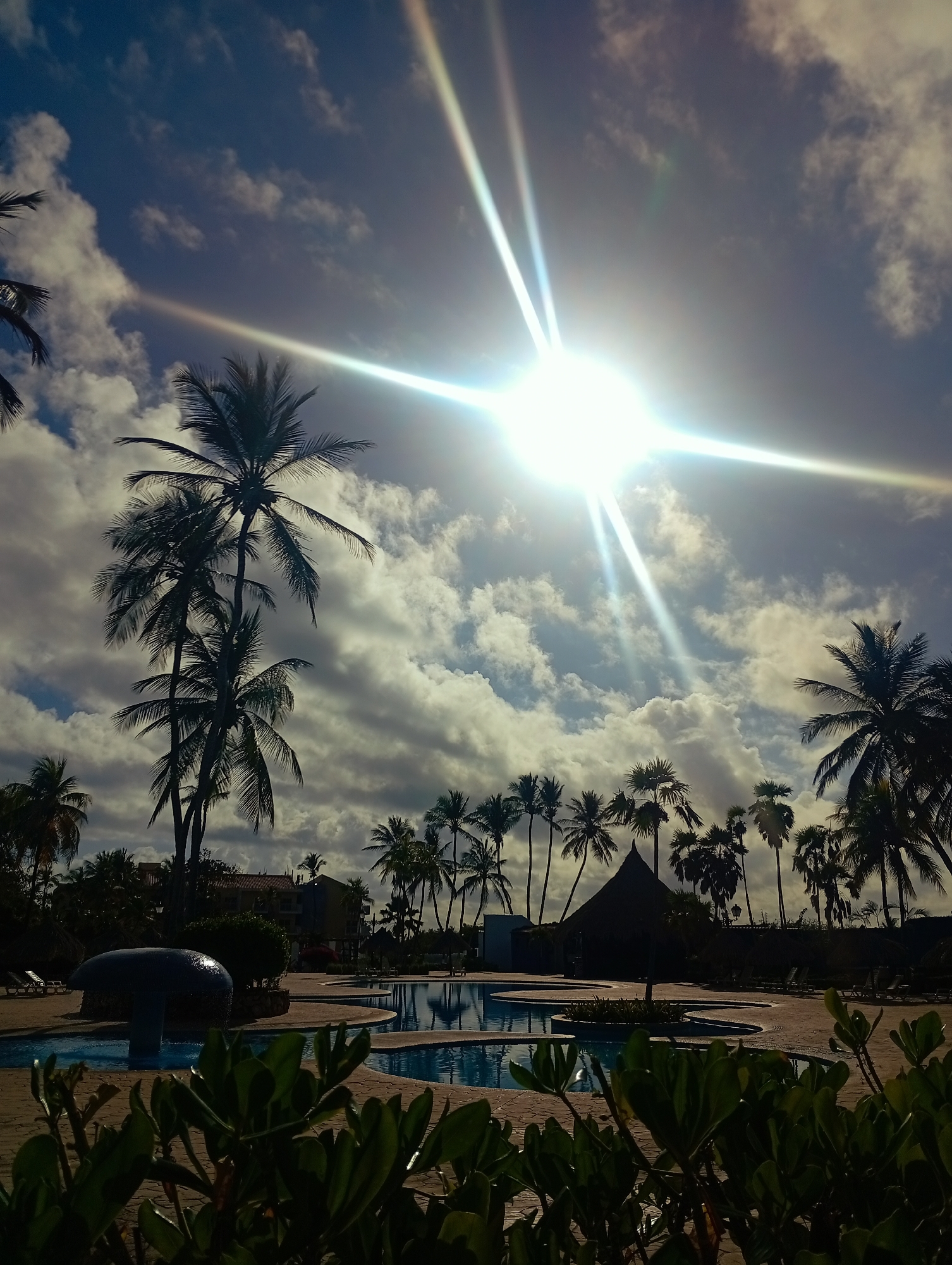 This image depicts an outdoor resort area in Playa El Agua, Venezuela. The scene is illuminated by strong sunlight originating from the upper right, producing prominent lens flares across a partly cloudy blue sky. Numerous palm trees, predominantly silhouetted, populate the midground and background, forming a dense canopy and lining the horizon. In the immediate foreground, out-of-focus green foliage is visible. The central midground features an irregularly shaped swimming pool with blue water, bordered by light-toned paved decking. A dark, mushroom-shaped water feature is situated within the pool area on the left. To the right of the pool, a dark, conical thatched-roof structure is visible. Further back, a building with a reddish-brown roof is partially discernible through the palm trees. Multiple empty lounge chairs are arranged on the paved surfaces surrounding the pool. No individuals are present in the scene.