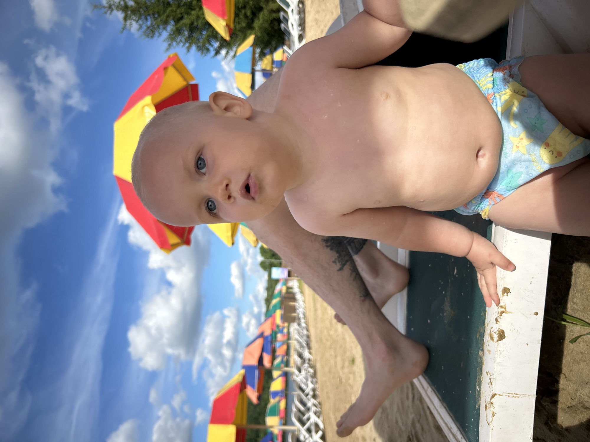 A close-up, low-angle shot shows a young child with blue eyes and fair skin, wearing swim trunks with a pattern of yellow starfish and other sea creatures. The child's mouth is slightly open, as if speaking or surprised. Part of an adult's legs, with visible tattoos on the shin, are also in the frame, suggesting they are holding or sitting next to the child.
The background is a bright blue sky with fluffy white clouds. Several colorful beach umbrellas with alternating red and yellow panels are visible, indicating a sunny outdoor setting. Some of these umbrellas are clustered together, and others are spread out further down what appears to be a sandy beach. In the distance, more umbrellas and some foliage can be seen. The child is positioned on what looks like the edge of a lounger or a similar piece of beach furniture.
The overall impression is one of a summer day at a beach resort, possibly in Юбилейный, Russia, given the location context. The lighting suggests it is daytime, likely late morning or early afternoon due to the bright sunlight casting shadows. The child's expression and the relaxed pose of the adult's legs convey a sense of leisure and enjoyment. No visible text is present in the image.