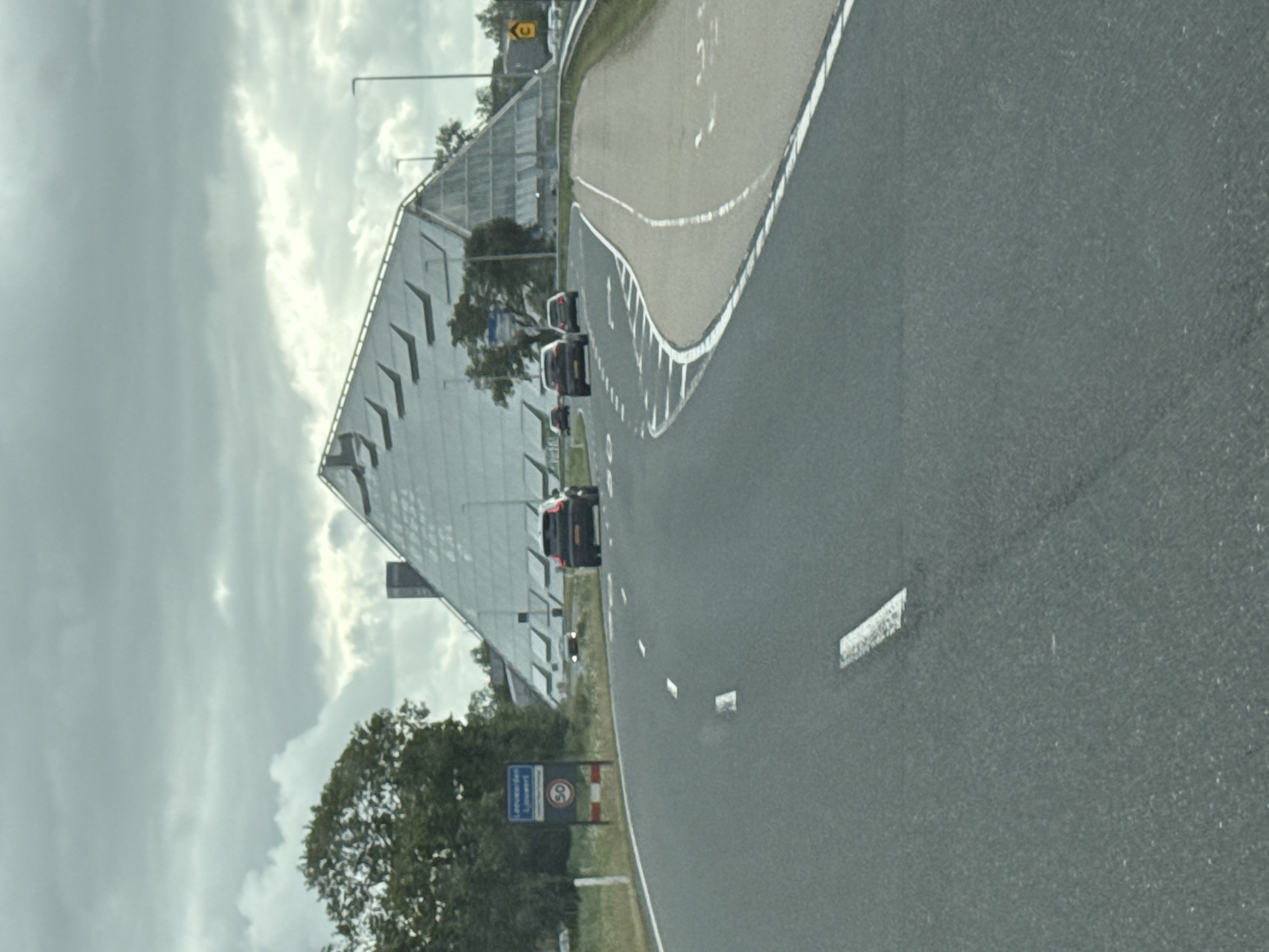 The image depicts a view from a road in Leeuwarden, Netherlands, approaching a distinctive modern building with a large, sloping, glass-paneled roof. The building resembles a geometric pyramid or a large tent. Several cars are visible on the road, some driving away from the camera. A blue sign with white text and a red circle containing the number "50" is posted on the left side of the road, indicating a speed limit. The sky is overcast with heavy clouds. The road is paved, with white lane markings visible. Trees are present on the left side of the road, with the building situated further in the background. The overall scene suggests an urban or suburban setting with a significant architectural landmark.