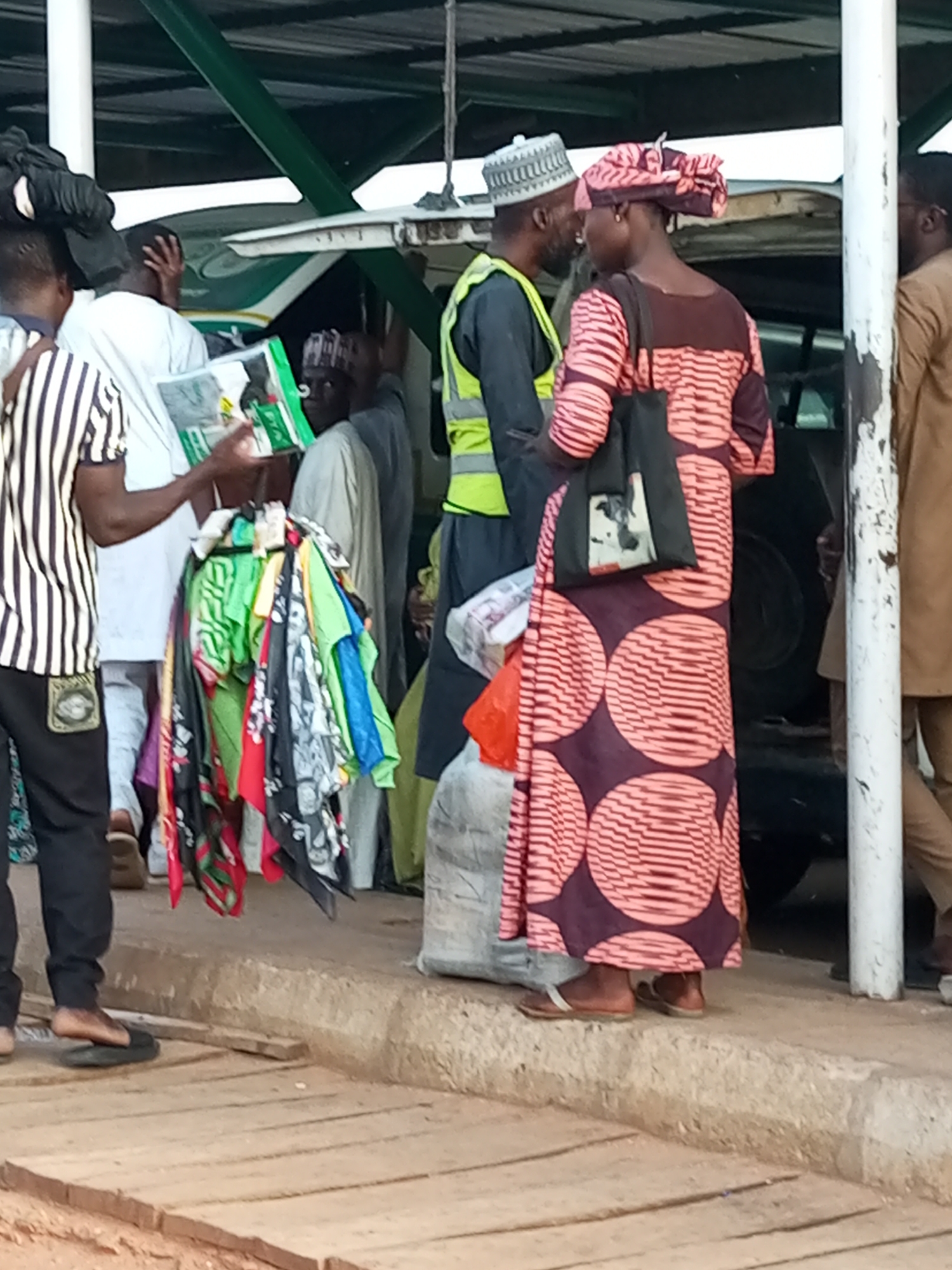 This image captures a scene at a bus park in Gombe, Nigeria, likely during a period of transit or waiting. In the foreground, a group of individuals are gathered, some appearing to be passengers and others possibly vendors or porters. A person on the far left, partially visible, is wearing a black and white striped shirt and black pants. To their right, another individual in a white shirt and light-colored pants holds a green package. In the center, a person wearing a yellow safety vest and a traditional cap is interacting with another person, a woman, who is dressed in a pink and maroon patterned garment, including a matching headscarf. She carries a black bag with a picture on it. A rack of colorful fabrics, appearing to be scarves or small garments, is displayed between them. Behind this group, various vehicles, including what looks like a bus, are present, partially obscured by the people and the covered waiting area. The ground is a mix of concrete and wooden planks. The overall atmosphere suggests a busy, everyday occurrence at a transportation hub.
