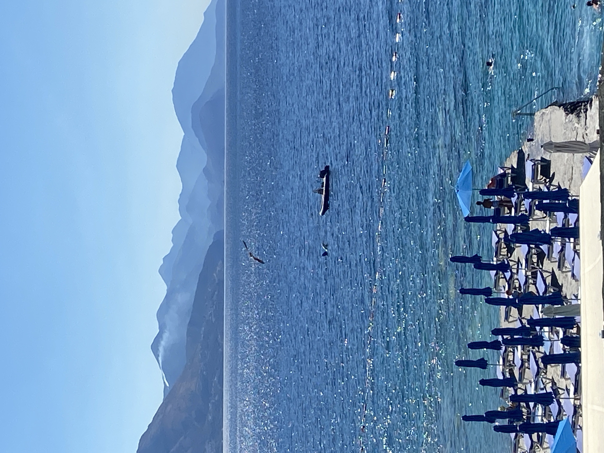 The image depicts a coastal scene with a body of water, mountains in the background, and a beach area in the foreground. The sea is a vibrant blue, with sunlight glinting off its surface. In the distance, a chain of mountains stretches across the horizon, with some atmospheric haze obscuring the farthest peaks. There is a small inflatable boat with two people on board in the middle of the water, and a bird in flight above it. Further to the right, a string of buoys marks a section of the water.
The foreground shows a beach with numerous rows of closed blue umbrellas and loungers set up on the sand. A concrete pier extends into the water, with a few people visible on it and in the sea. One person is standing under a bright blue umbrella on the pier, while another is partially reclined on a lounger. Two people are swimming in the sea near the pier. The overall impression is a sunny day at a resort or beach area. The scene is located in Prijevor, Montenegro.