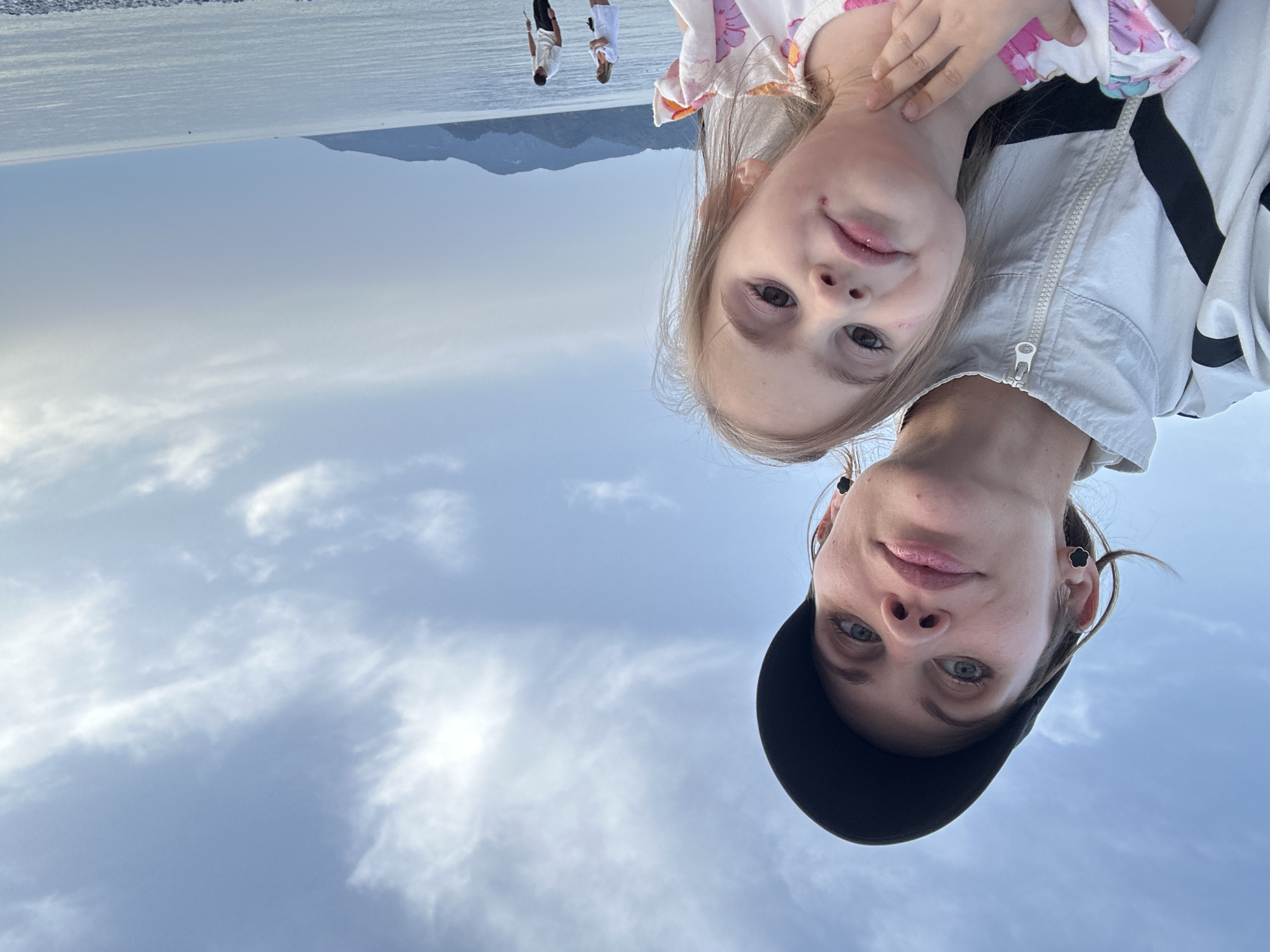 The image shows a woman and a young girl, possibly related, facing the camera. They appear to be outdoors, with a body of water and mountains visible in the background. Other people are standing in the distance. The sky is cloudy. The photograph is oriented upside down. Although the location is given as Fethiye, Turkey, more information is needed to confirm this.