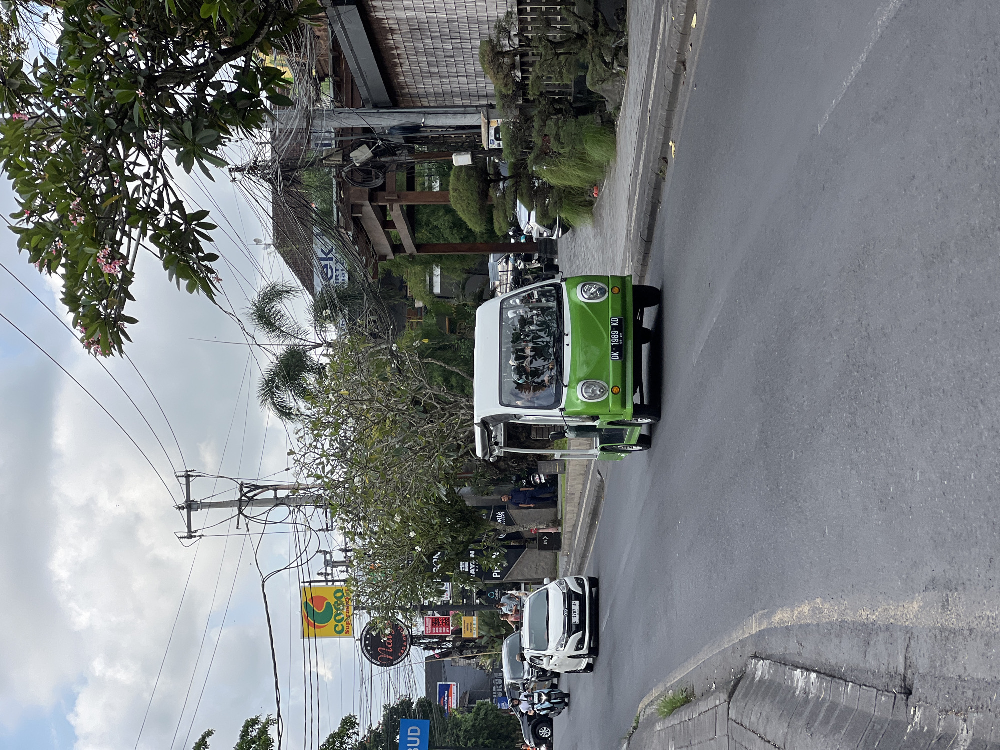 The image depicts a street in the Ubud District, Indonesia, where a three-wheeled green and white motor vehicle bearing the license plate "DK 1989 KD" is shown driving along the road. A white car with the license plate "L 1787 AF" is also visible, parked on the roadside. A sign for "COCO Supermarket" is present alongside various other commercial signage and businesses. Vegetation including trees and greenery is visible. There is a utility pole with numerous wires. People can be observed on the sidewalk. The sky is overcast. The road surface is grey asphalt.