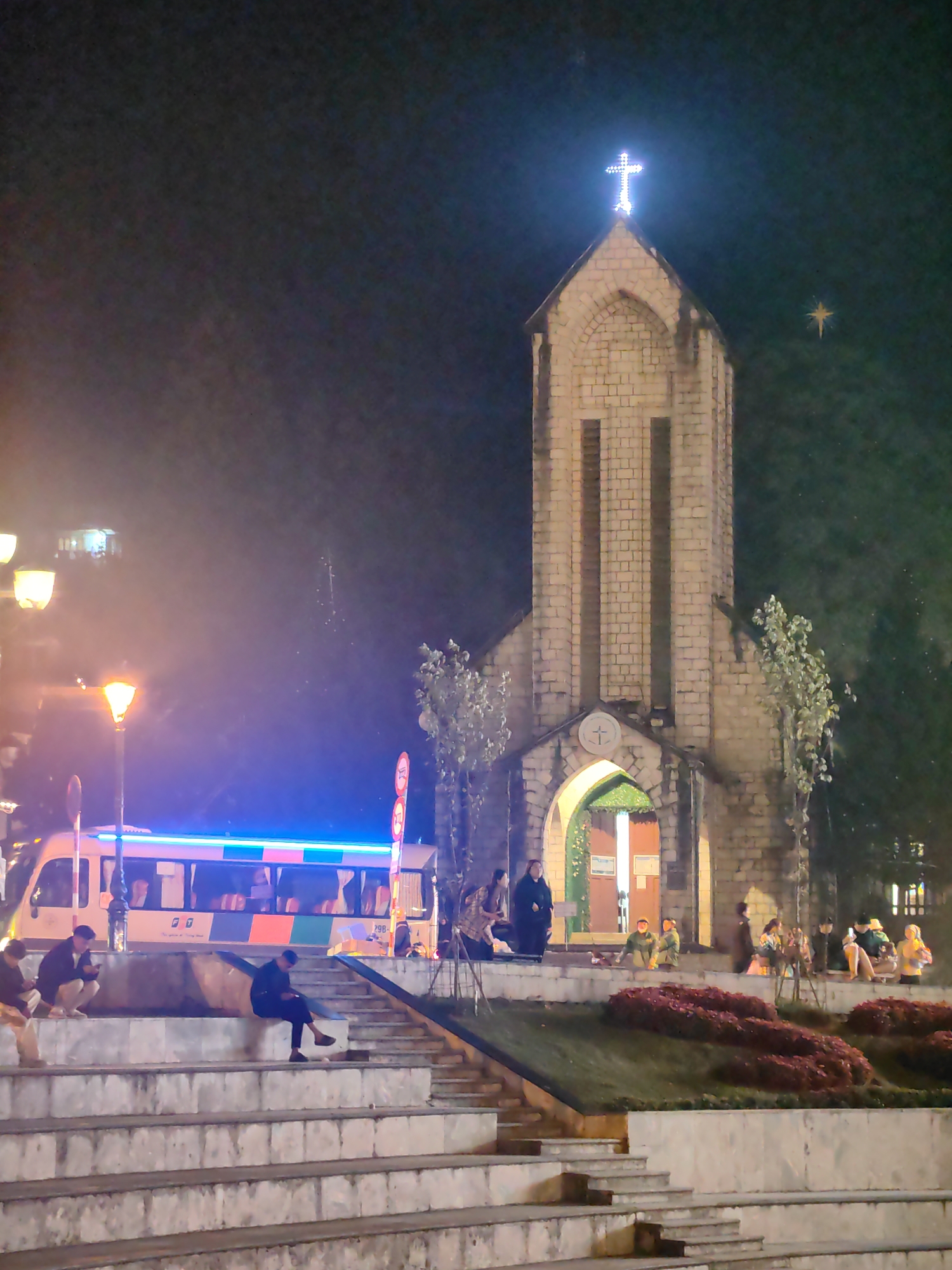 The image depicts a nighttime scene centered on the Sa Pa Stone Church in Sa Pa, Vietnam. The church, a Romanesque revival structure built from light-colored stone blocks, stands prominently in the central-right, topped by a brightly illuminated white cross. Its main arched entrance is visible and lit from within.
In the foreground and midground, a wide set of multi-tiered, light-colored concrete or stone steps descends from the left. Several individuals are present: three men are seated on the steps on the left, two appearing to interact with mobile devices. Another man sits alone on a higher step to the right, also focused on a mobile device.
To the left of the steps, a white passenger bus with horizontal blue, green, and orange stripes is parked, featuring a continuous blue LED light strip along its roofline. Two amber-glowing streetlights illuminate this area. A red circular traffic sign with a white bar is partially visible near the bus.
In front of the church, a sloped grassy area features neatly arranged flowerbeds of dark red or purple foliage. Numerous individuals are gathered on the church grounds and near its entrance, standing and sitting, appearing to socialize. Small trees with light green leaves are visible around the church.
The dark night sky, with atmospheric haze, forms the background. A distinct, glowing yellow star-shaped decoration is visible in the upper right quadrant of the sky, separate from the church. Faint artificial lights from other buildings are discernible in the distant background on the left.