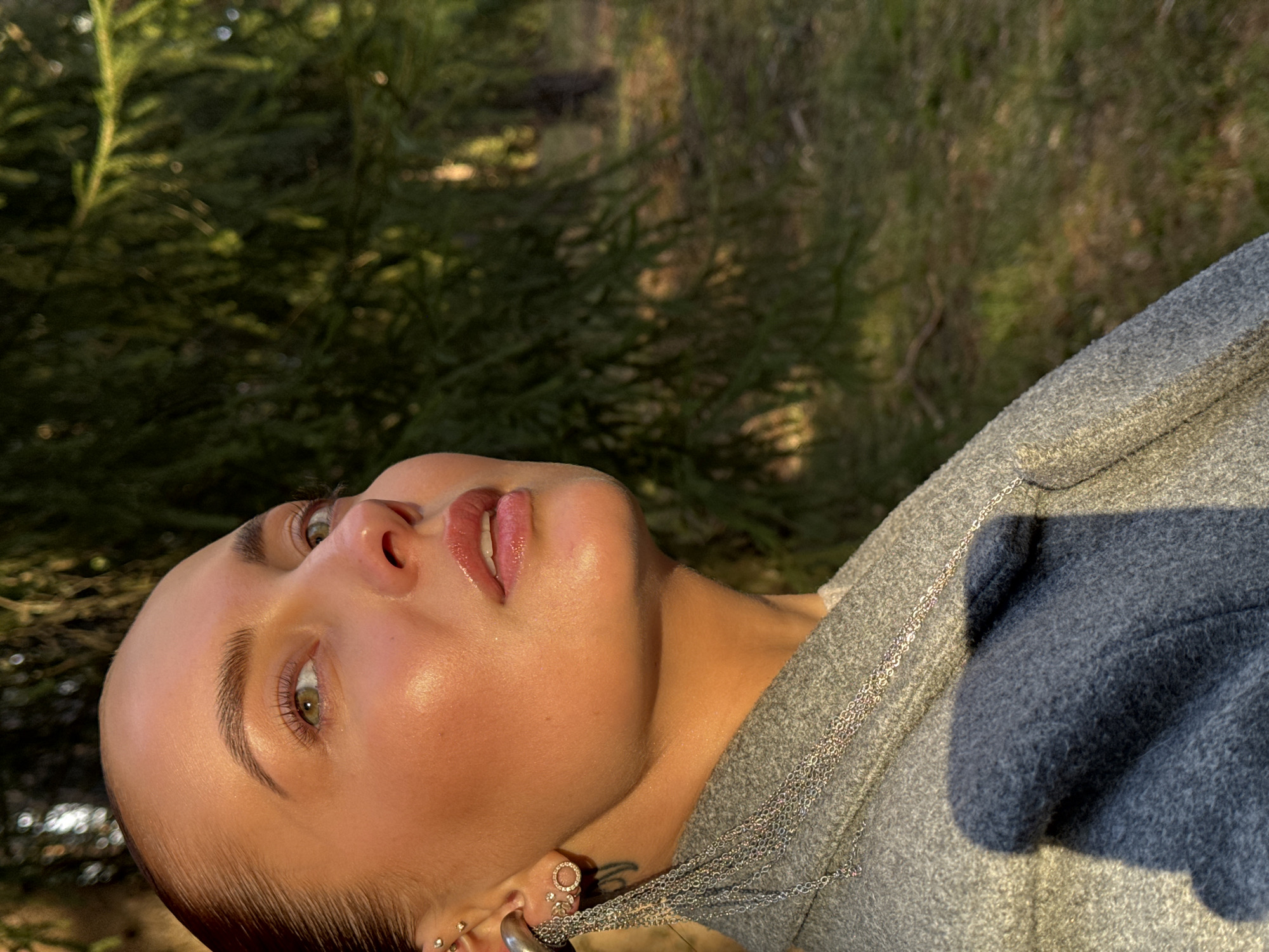 A close-up portrait shows a young woman looking upwards, her head tilted towards the right side of the frame. She is positioned against a blurred background of green foliage, suggesting an outdoor, natural setting like a forest or wooded area in Прикамский, Russia.
The woman's face is illuminated by warm, golden sunlight, indicating either early morning or late afternoon. Her skin appears dewy and highlighted, particularly on her cheeks, forehead, and lips. She has striking green or hazel eyes looking towards the upper left, prominent, well-groomed eyebrows, and full, glossy, slightly parted lips. Her hair, which appears dark, is pulled back from her face.
She is wearing a textured, grey garment, possibly a sweater or jacket. Multiple thin silver chain necklaces are draped around her neck and over her clothing. In her visible left ear, she wears several earrings, including a small hoop, various studs, and a larger, circular earring. A small tattoo is faintly visible on her neck or upper shoulder area, partially obscured by the chains. The overall impression is a serene and artistic portrait taken in natural light. There is no visible text in the image.