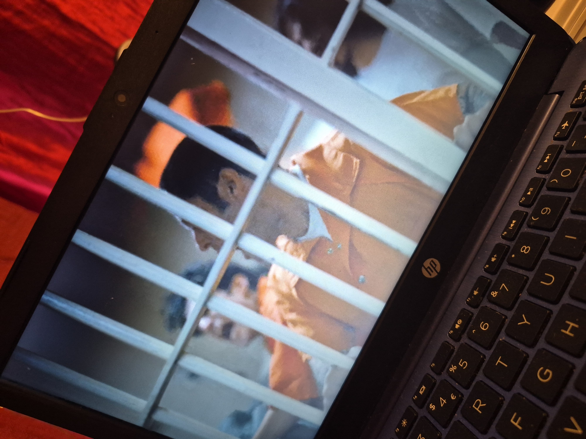 A laptop displays a scene from what appears to be a prison cell, with a person in an orange jumpsuit visible behind bars. The laptop keyboard is in the foreground, with several keys visible, including "R", "F", "G", "V", "T", "Y", "U", "H", "6", "7", "8", "9", and "0". The laptop screen is reflecting some of the environment around it. The background of the laptop screen is a blurred image of a person in an orange jumpsuit, with a mustache, behind light-colored bars. The environment outside the laptop is a red textured surface. The image does not provide any information about the location in the city of Sandwell, United Kingdom.
