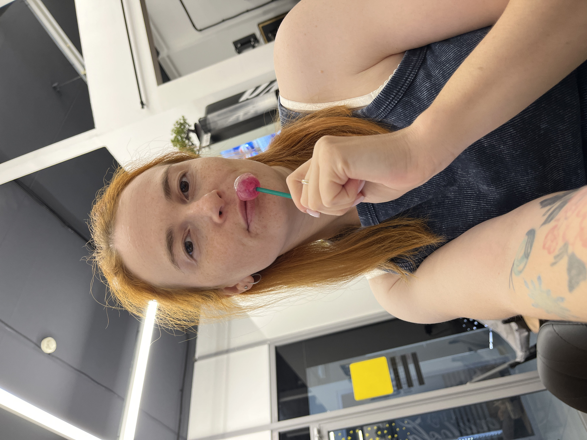 A woman with red hair is holding a pink lollipop to her lips and looking at the camera. She is wearing a dark blue ribbed tank top and a silver ring. Her left arm, which is visible, has a tattoo of flowers and leaves. The background appears to be an indoor space, possibly a salon or a commercial establishment, with white walls, ceiling lights, and large windows. A yellow rectangular sign is visible on the window to the left. The overall lighting is bright. No specific location in Chaikovsky, Russia, can be identified from the image.