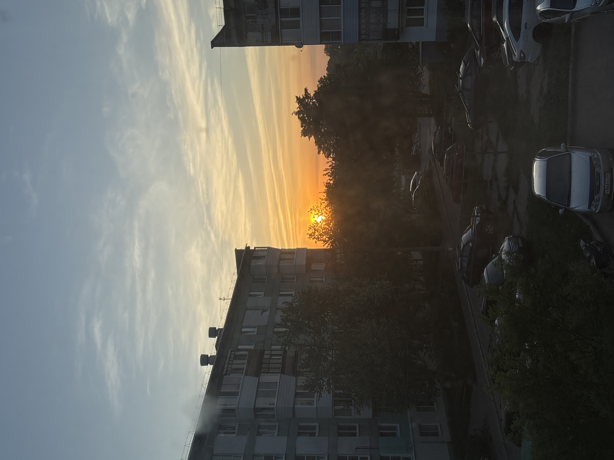 View from a high angle looking down toward the ground showing the sky near sunset in the city of Chaikovsky, Russia. Buildings stand on the left and right. Trees block the view to the orange sky. Cars are parked on the street along with trees.