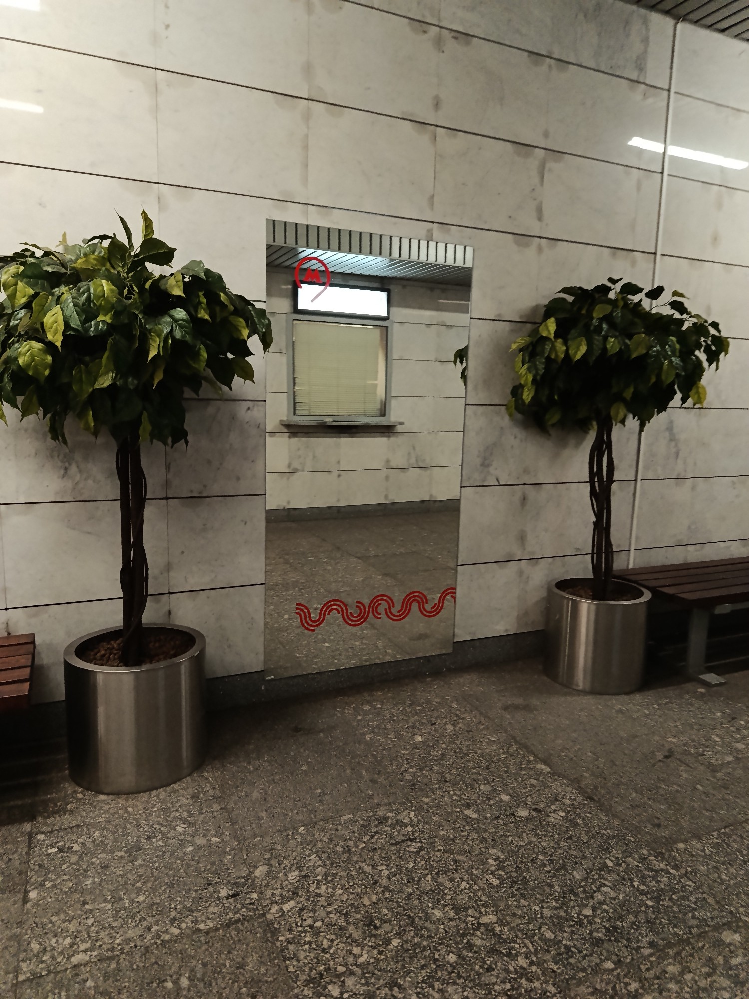 In an indoor environment with a marble-tiled wall and a speckled granite floor, two artificial potted trees flank a large mirror. The tree on the left is positioned to the left of the mirror, and the tree on the right is to the right. The mirror reflects an opening with a red circular sign that contains a white letter "M" and a horizontal line, possibly indicating a metro station. Red wavy lines are visible on the bottom portion of the mirror. A wooden bench is partially visible to the far right of the frame. The overall scene appears to be in a public space, possibly a train station or a subway platform.