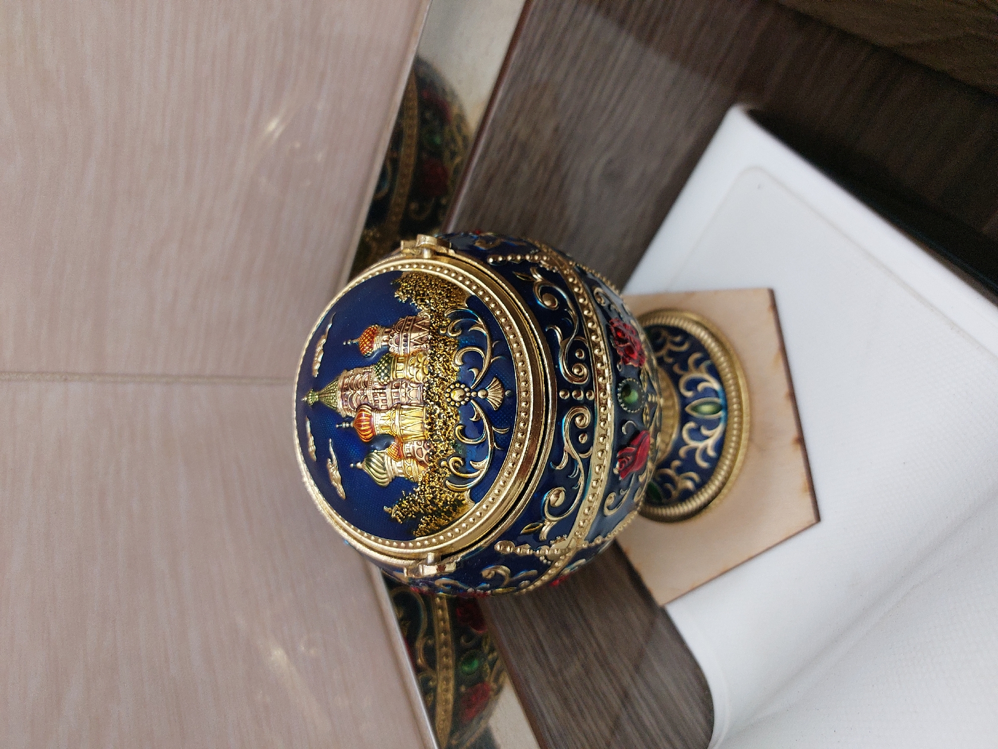 A darkly ornate, spherical object, possibly an ashtray or decorative box, rests on a white surface in a corner. The object is predominantly dark blue with intricate gold embellishments and features a detailed depiction of a multi-domed building, likely St. Basil's Cathedral in Moscow. The background consists of light-colored tiled walls and dark wood paneling. No people or actions are depicted, and the image appears to be a still life. There is no information to indicate the location in the city of Nakhabino, Russia.