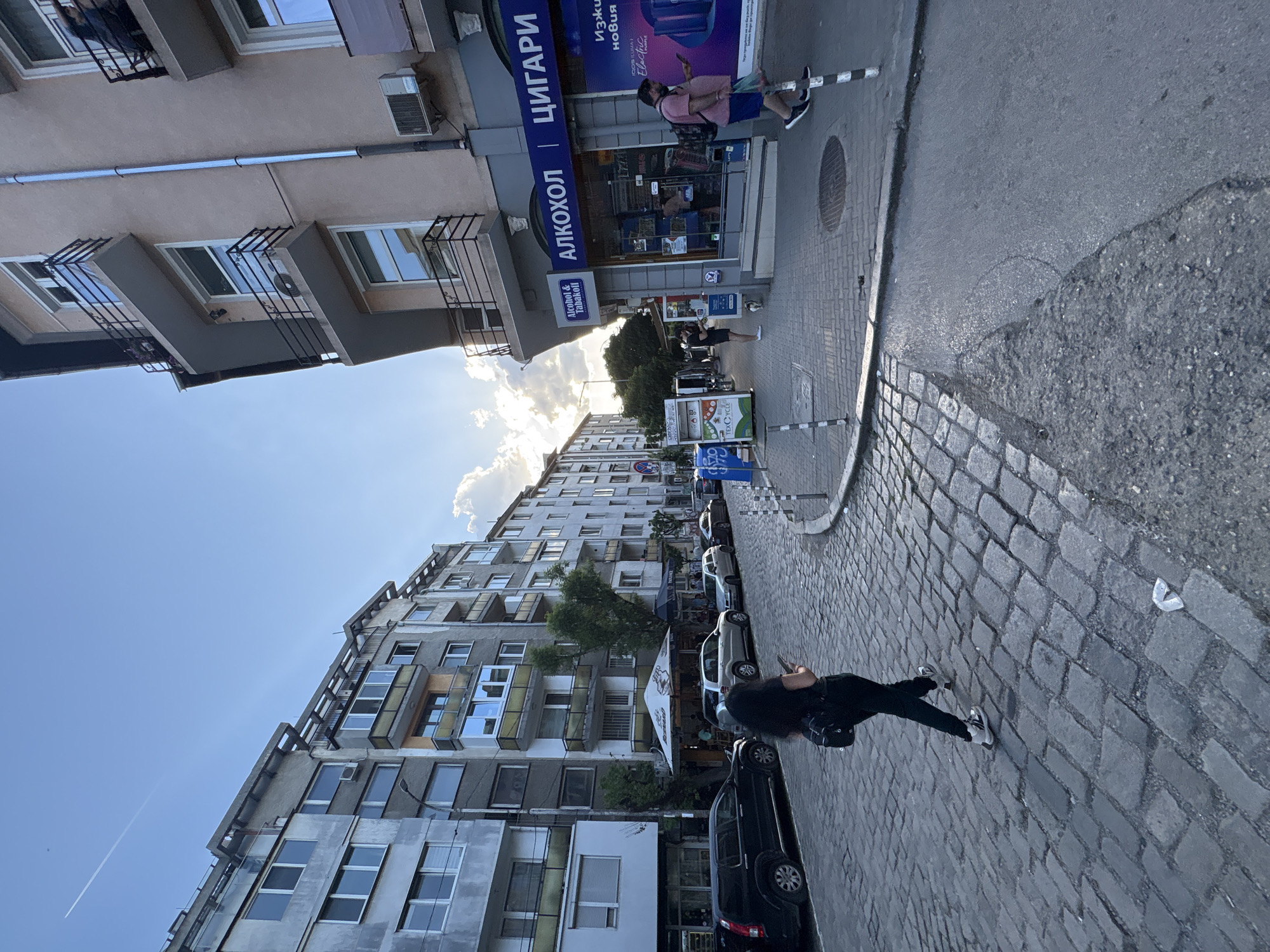 This image captures a street scene in Sofia, Bulgaria. It shows buildings of varying heights and architectural styles, typical of urban landscapes. The sky is partly cloudy with the sun shining through. Street-level shops and advertisements are visible, with signs in Cyrillic script. A few cars are parked along the side of the street. Two individuals are present on the sidewalk; one is walking near a shop, while the other is standing on the cobblestone walkway, appearing to use a mobile phone.