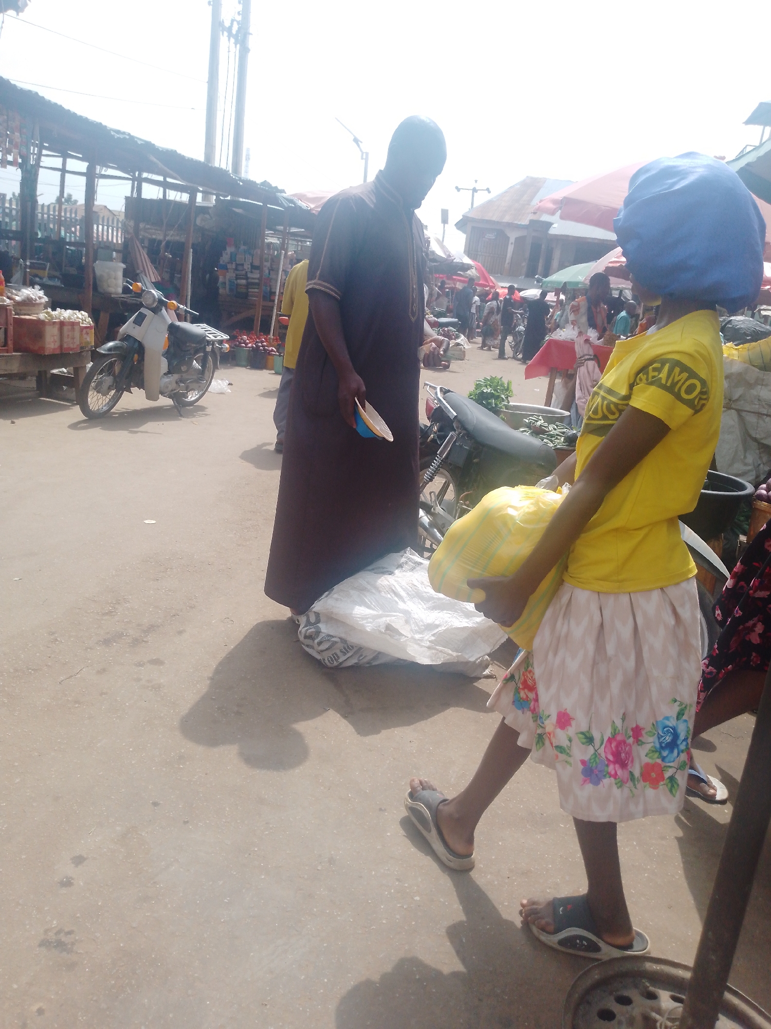 In an outdoor market in an urban setting, a person wearing a long brown robe stands next to a large white bag. A child in a yellow t-shirt and floral skirt carries a yellow bag. A motorcycle is parked nearby. Other people and vendor stalls are visible in the background.