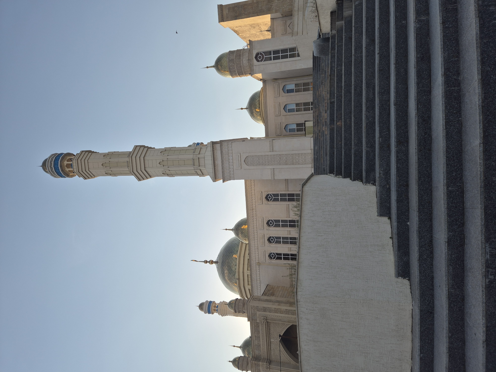 The image shows a portion of the Nauryz Mosque in Kokshetau, Kazakhstan. Visible are the minaret extending into a clear blue sky and the mosque's domes and upper stories. To the right, a section of a stone wall or staircase runs vertically along the image. A bird is flying in the upper part of the sky.