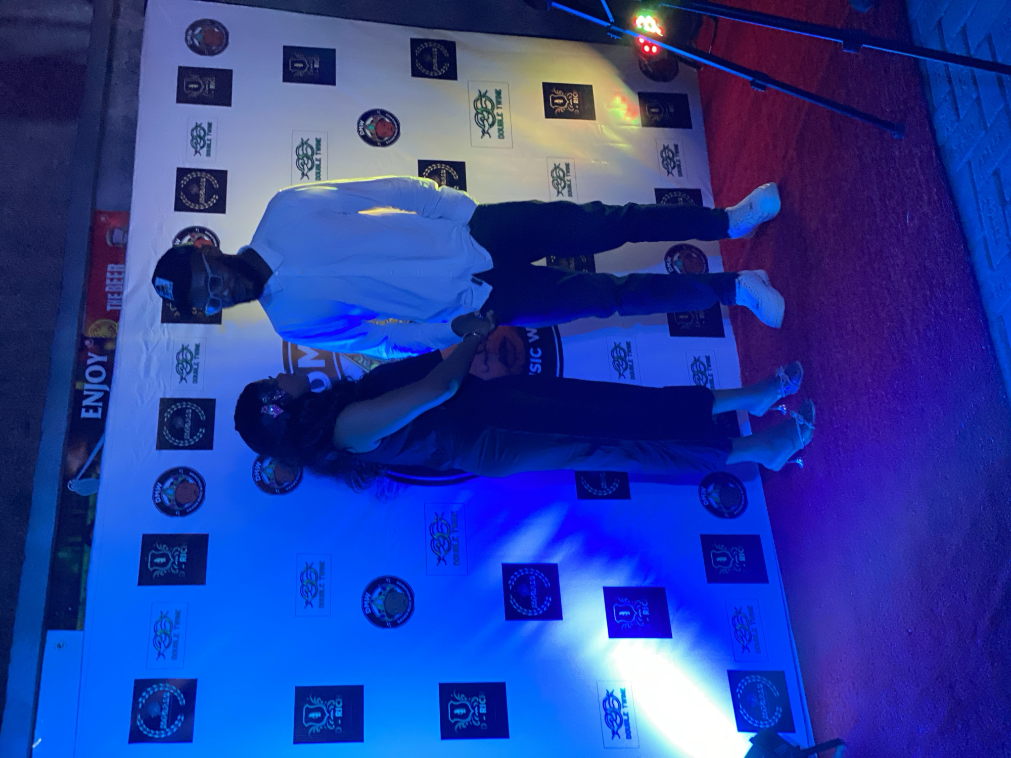 The photograph depicts a man and a woman posing in front of a backdrop. The man is wearing a white shirt, dark pants, a hat, and white shoes. The woman is wearing dark clothing. The backdrop features various logos and text. The scene is illuminated by blue lighting. The location is inferred to be Iranla, Nigeria based on the context.