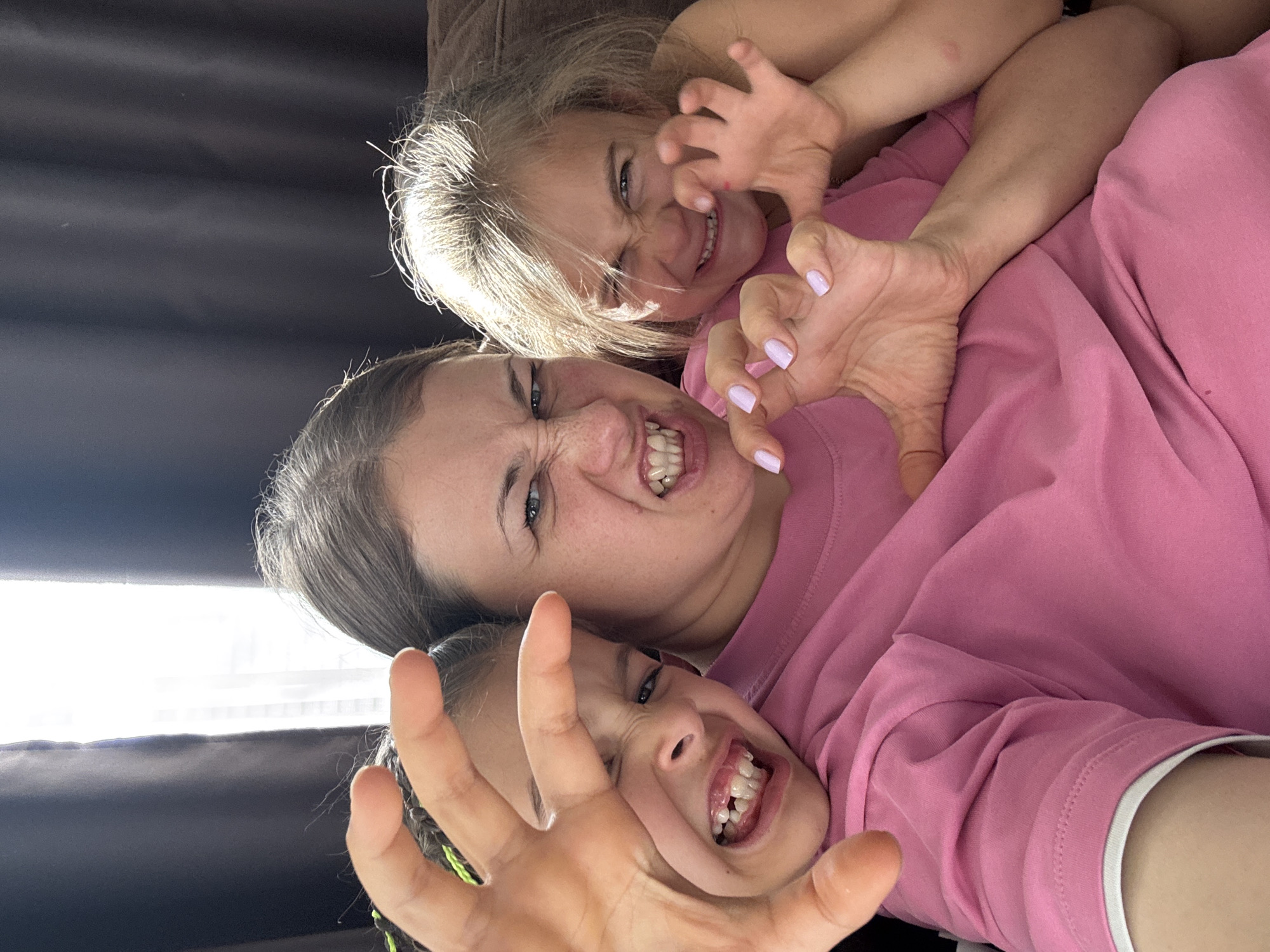 A medium shot photo shows three people, likely a mother and two daughters, wearing pink shirts. They are making fierce facial expressions and clawing gestures with their hands, appearing to be playing a game. The background consists of dark curtains and a bright window, suggesting an indoor setting, possibly a home, in a city like Uyutnoye, Ukraine. The lighting is natural and comes from the window.
