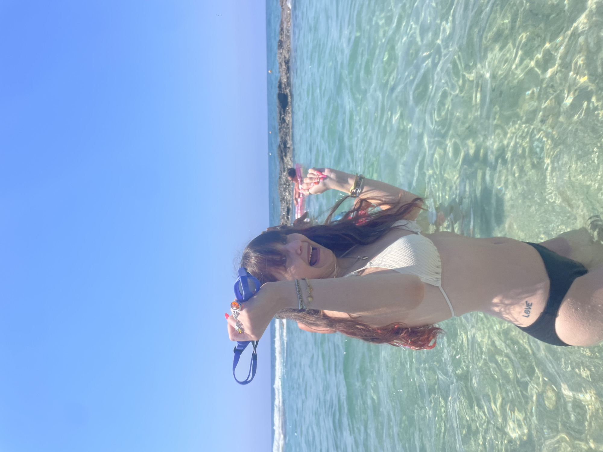A young woman is standing in shallow, clear ocean water, with her mouth open in a surprised or excited expression. She is wearing a white bikini top and black bikini bottoms. She has long, dark, wavy hair with some reddish-pink highlights. On her left hand, she is holding a pair of blue swimming goggles above her head, and she is wearing several rings and bracelets on her arms. A tattoo of the word "love" is visible on her left hip. The water is clear enough to see the sandy bottom. In the background, the ocean stretches out to the horizon under a clear blue sky. Waves are breaking in the distance, and there are rocky formations in the water. Another person can be seen further back in the water, floating on a pink inflatable. The scene appears to be a beach in Paphos Municipality, Cyprus, on a sunny day.