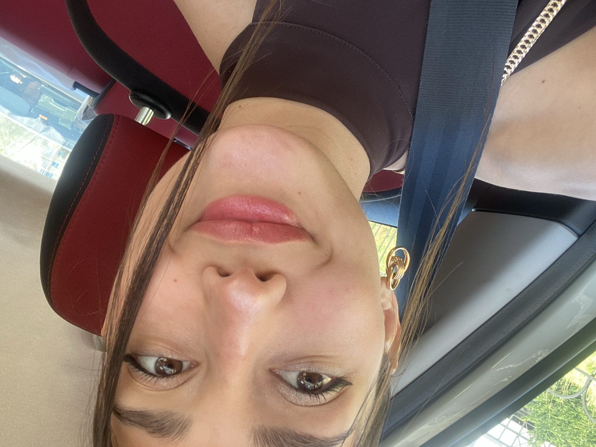A woman is wearing a brown shirt and a blue seatbelt in a car. She has brown hair and gold hoop earrings. The interior of the car is red and black. The location may be in Budva, Montenegro, but there are not enough clues to determine this location for sure.