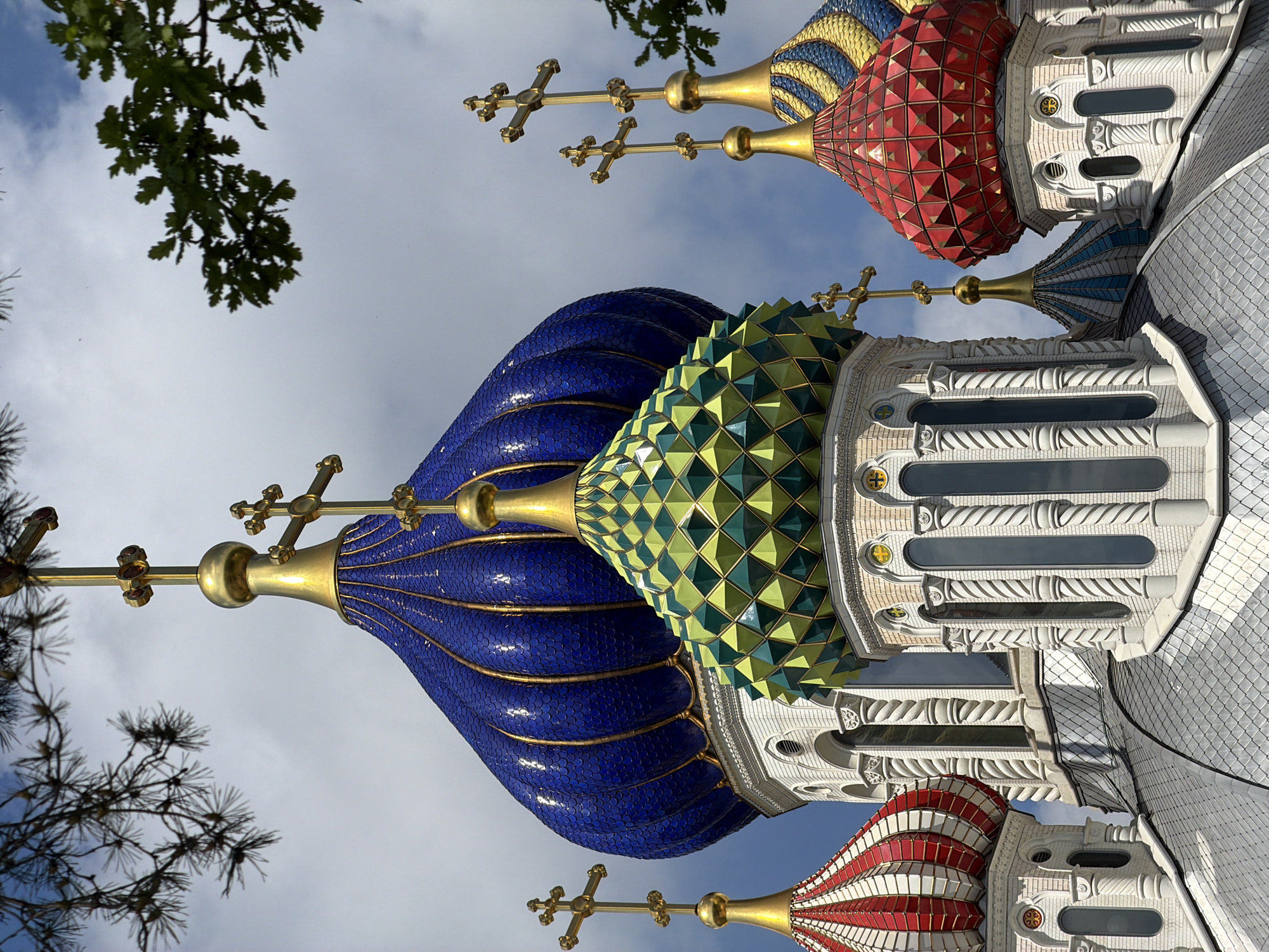 The image shows a low-angle view of the colorful domes of Saint Basil's Cathedral in Moscow, Russia, against a cloudy sky. The domes are adorned with intricate patterns and various colors, including blue, green, and red. Golden crosses top each dome. Tree branches partially frame the view. The architectural details of the cathedral's exterior are visible, including the white walls and decorative elements.