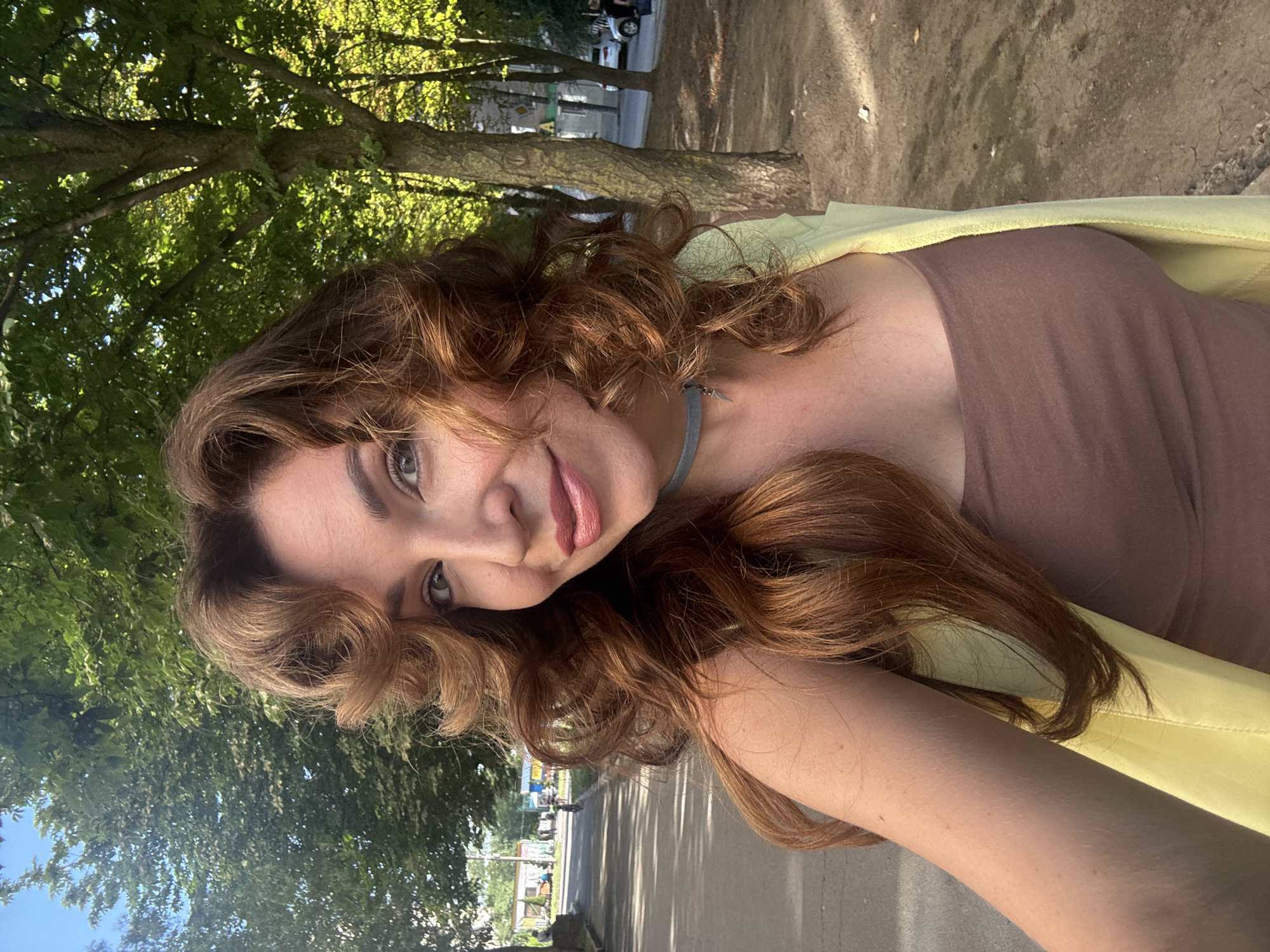 A medium shot photo captures a woman with auburn, wavy hair and light makeup posing outdoors. She is wearing a brown tank top and a yellow cardigan. A thin, light blue choker with a small pendant rests on her neck. She is smiling and looking directly at the camera. The background suggests a park or tree-lined path with a road visible to the left, indicating an urban environment. Based on the context provided, this scene is located in the city of Kryvyi Rih, Ukraine. The lighting indicates it is daytime, likely late afternoon due to the shadows cast by the trees. Other people and vehicles are distantly visible in the background, suggesting a public space.