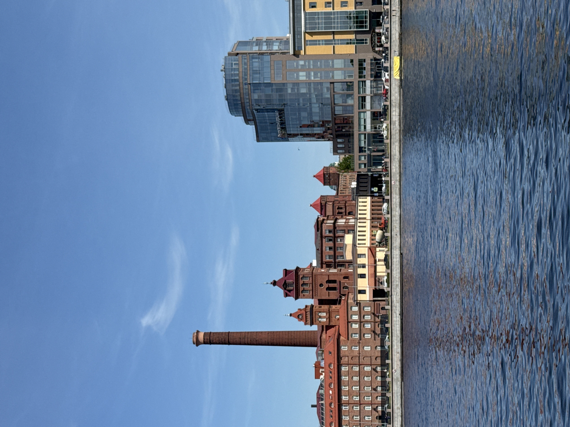 The image shows a view in Saint Petersburg, Russia. On the left side, there's a clear blue sky. Along the shoreline, there are various buildings, including a tall brick chimney and several brick buildings with red roofs. Further back, there is a modern glass and yellow building. On the right side, the image shows the surface of a body of water, possibly a river or canal, with visible ripples on the water.