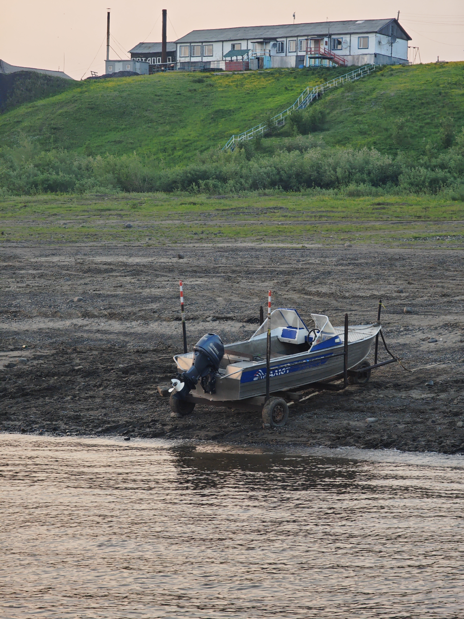 This is a forensic, matter-of-fact description of the provided image, which depicts a scene in Potapovo, Russia.
The image showcases a small, aluminum motorboat resting on a trailer on a gravelly shore. The boat is a light silver color with blue accents, featuring a dark blue outboard motor labeled "YAMAHA" on the stern. The interior of the boat has blue seating and a steering console. The trailer has two wheels on each side and is positioned on the shore, which is composed of mud, gravel, and scattered rocks.
Behind the boat, the shoreline extends towards a grassy hill. The hill is covered in green vegetation and dotted with small yellow flowers. On top of the hill sits a long, white, multi-story building with a gray roof. The building has multiple windows and doors, and a red-railed staircase leads up the hill to the building's entrance. Adjacent to the building are two tall, dark chimneys, suggesting industrial or heating facilities.
The foreground of the image shows a body of water with ripples reflecting the soft, possibly evening, light. The water appears calm, with subtle golden and pink hues. The environment suggests a rural or semi-industrial setting near a body of water, consistent with a location in Potapovo, Russia. No people are visible in the image.