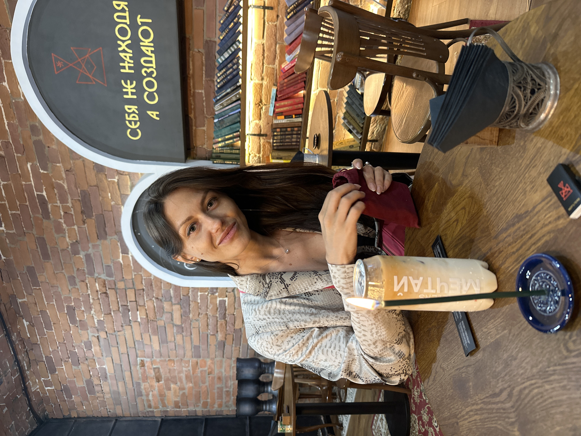 A woman with long dark hair and brown eyes, wearing a snakeskin-patterned blazer over a dark top and a pink crossbody bag, is seated at a wooden table in what appears to be a cafe or restaurant. She is looking towards the camera with a slight smile, holding a small dark red pouch in her hands. In front of her, a clear plastic bottle with a light orange beverage and Russian text that reads "МЕЧТАЙ" (Dream) is lit with an incense stick. A dark blue incense holder sits on the table next to the bottle. Also on the table are black napkins in a vintage silver napkin holder, a small black object with a red symbol, and what appears to be a menu or card. The background shows exposed brick walls, shelves filled with books, and wooden chairs. The overall ambiance suggests an indoor, possibly cozy, setting. The text on the wall behind her reads "СЕБЯ НЕ НАХОДЯ А СОЗДАЮТ," which translates to "They do not find themselves but create themselves." This phrase, combined with the cafe setting and the symbol on the wall and small object, could indicate the location is a creative space or a place that promotes self-expression, common in areas like the creative cluster in Saint Petersburg, Russia.