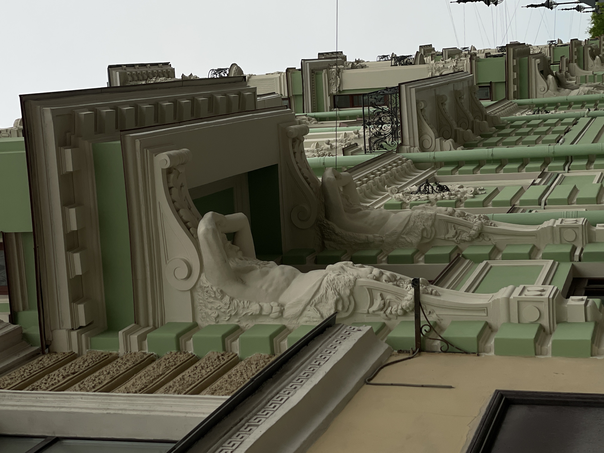 The image shows a close-up, low-angle view of the architectural details of a building, specifically the facade, likely located in Saint Petersburg, Russia, based on the style. The building features a pale green and white color scheme. A prominent architectural element is a series of reclining human figures, possibly statues or sculpted reliefs, positioned horizontally along the building's facade. The figures are classical in style, with draped clothing and intricate detailing. The architectural elements are arranged in repeating patterns, creating a sense of rhythm and order. The sky is overcast.
