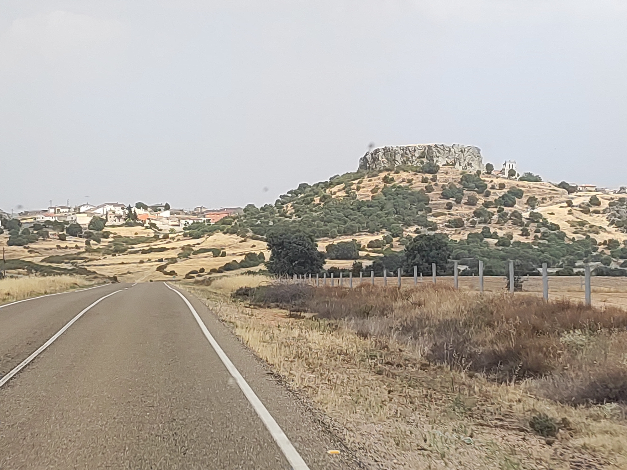 The image shows a rural landscape with a road leading towards a hill. The hill is topped with rocky formations and some structures, possibly ruins or a small building. A village with white- and red-roofed houses is visible on the left side of the hill. The foreground features a paved road with white lines, bordered by dry grass and vegetation. A fence runs along the right side of the road. The sky is overcast and light. The scene appears to be in a dry, Mediterranean climate. The overall impression is of a historical or ancient site within a rural setting. Based on the provided context, the location is Peñausende, Spain.