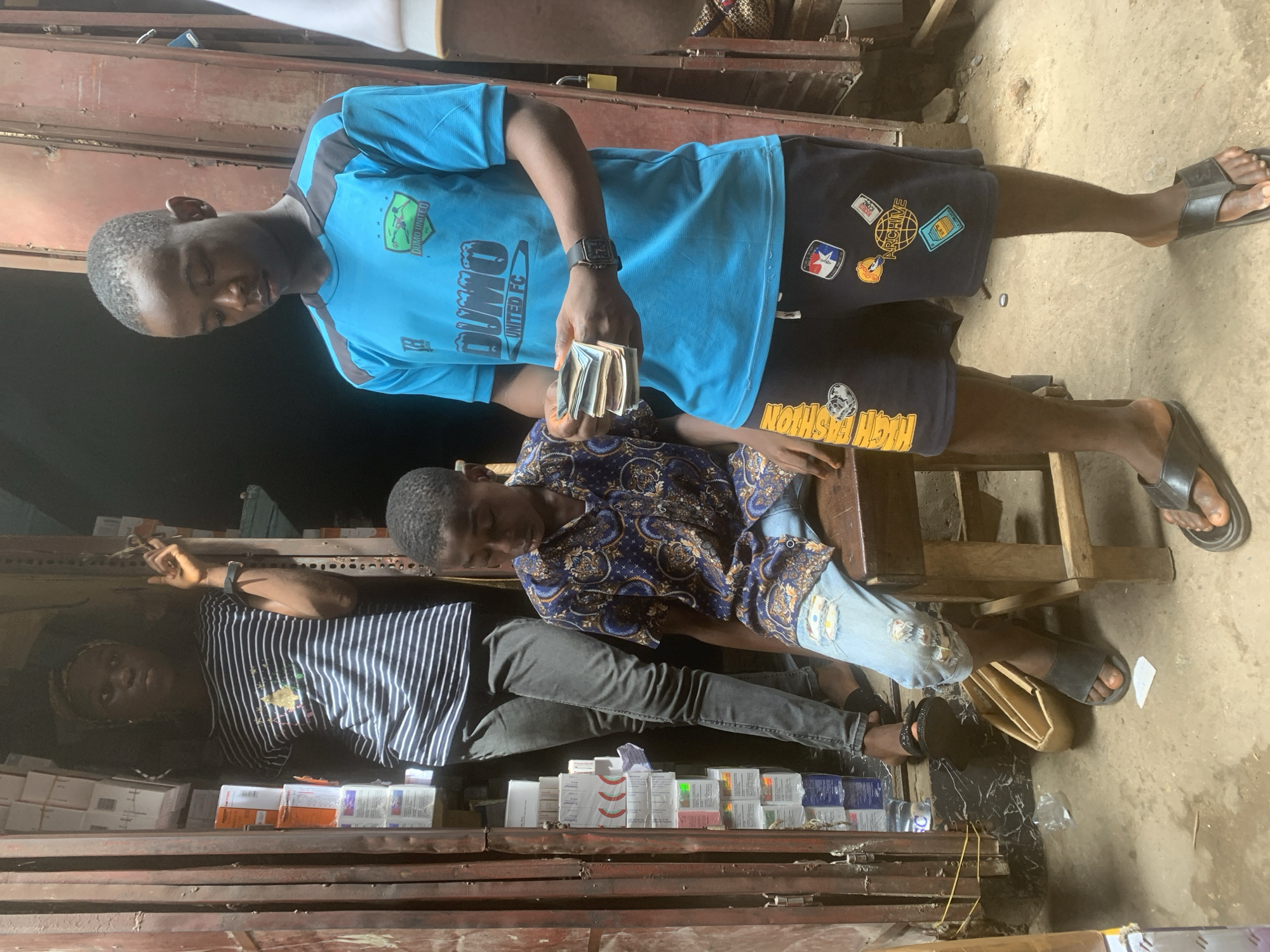 This image, set in Fegge, Nigeria, depicts three young men in what appears to be an informal market stall or a small shop.
In the central foreground, a young man stands, holding a thick stack of banknotes, likely Nigerian Naira, in his right hand. He is wearing a light blue t-shirt emblazoned with "DUMO UNITED FC" and black shorts featuring various patches, including one that reads "ARCHIVE" and another partially visible "HIGH FASHION" text. He has a dark wristwatch on his left wrist and wears black flip-flops. His attention is focused downward, presumably on the money.
To his right, another young man is seated on a wooden stool, leaning back with his eyes closed or downcast, indicating a moment of rest. He wears a blue long-sleeved shirt with a distinctive pattern and light blue ripped jeans, also paired with dark flip-flops.
Further to the left and slightly behind, a third individual is seen lying down or reclining on a surface, wearing a striped t-shirt and dark pants, possibly asleep or relaxing.
The immediate surroundings are filled with numerous small boxes and packages, stacked on what appears to be shelves or a counter made of rough wood or metal. Some of these packages are identifiable with the text "Celexat", suggesting they could be medication or health products. The background features simple wooden or corrugated metal structures, indicative of a modest commercial space. The ground is a mix of dirt and concrete, with a yellow cable visible. The natural lighting suggests it is daytime. The scene conveys a casual atmosphere, possibly during a transaction or a break from work.