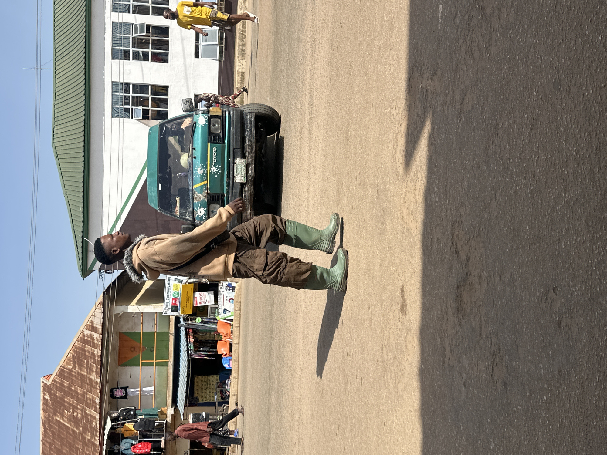 A man stands in the middle of a street in Bukuru, Nigeria. He is wearing a light brown jacket with a fur-lined collar, brown cargo pants, and tall, light green rubber boots. He is looking upwards and to his left. Behind him, a green Toyota van is parked on the side of the road. The van has "TOYOTA" written on its front, along with some decorative white splatters and a license plate. Several people are visible near the van, and further back, a street market with stalls selling clothing can be seen. The buildings in the background appear to be made of concrete and metal sheeting. The sky is clear and blue, indicating a sunny day. The lighting suggests it is daytime. The overall scene depicts a busy street in a Nigerian town. Visible text includes "TOYOTA", "POS", and "Young Communications".