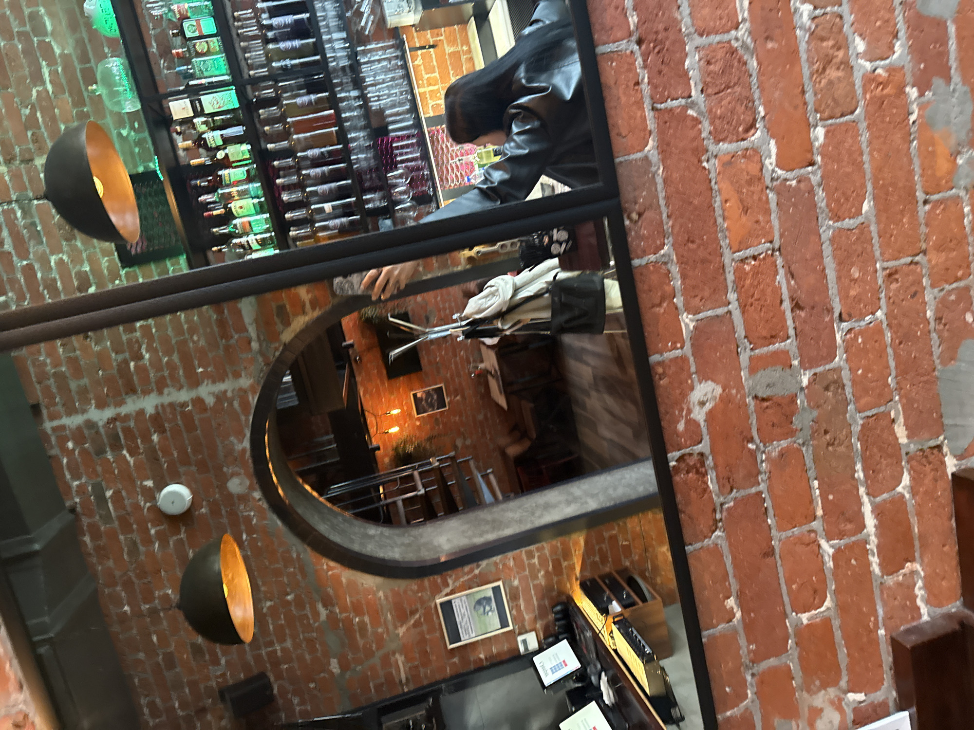 This image shows a view through a mirror inside a building. The interior features exposed brick walls. On one side, the mirror reflects a bar with a large selection of bottled beverages on display. In the foreground, a person wearing a black leather jacket is partially visible, leaning over the mirror. The mirror also reflects a lower level of the establishment with tables and chairs. Lighting fixtures, including spherical pendant lamps with a gold interior, are visible. The photo appears to be taken in a bar or restaurant setting. Location information is not ascertainable from the information given.