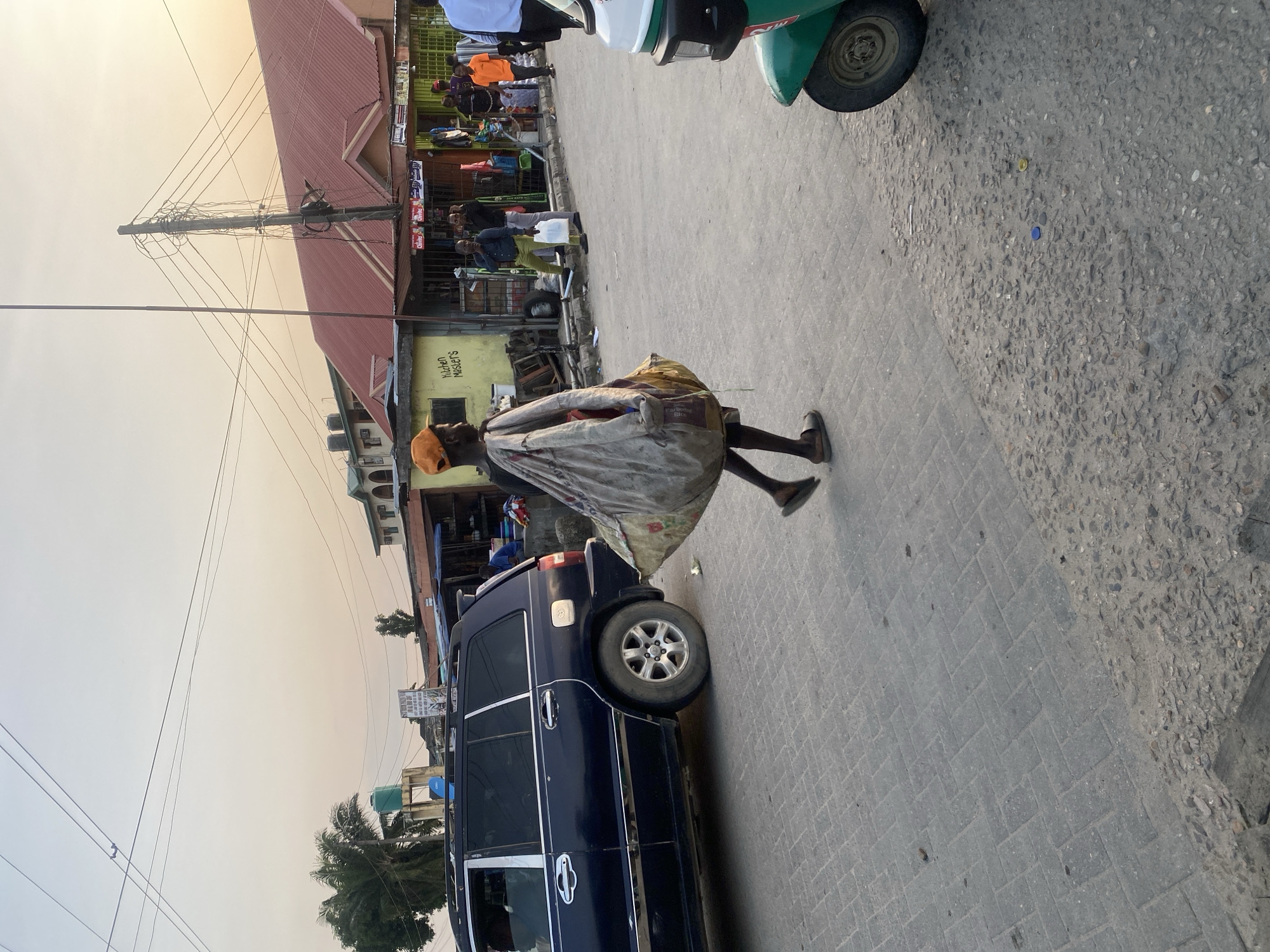 A man is walking on a paved road in Ado, Nigeria, carrying a large, bulky bag over his shoulder. He is wearing an orange cap and sandals. To his left, a dark blue SUV is parked on the road. In the background, there are several buildings with red roofs, some appearing to be shops or market stalls. A green and white tricycle is visible on the right side of the frame. Power lines are strung overhead, and palm trees are present in the distance. The sky is clear and appears to be late afternoon or early evening.