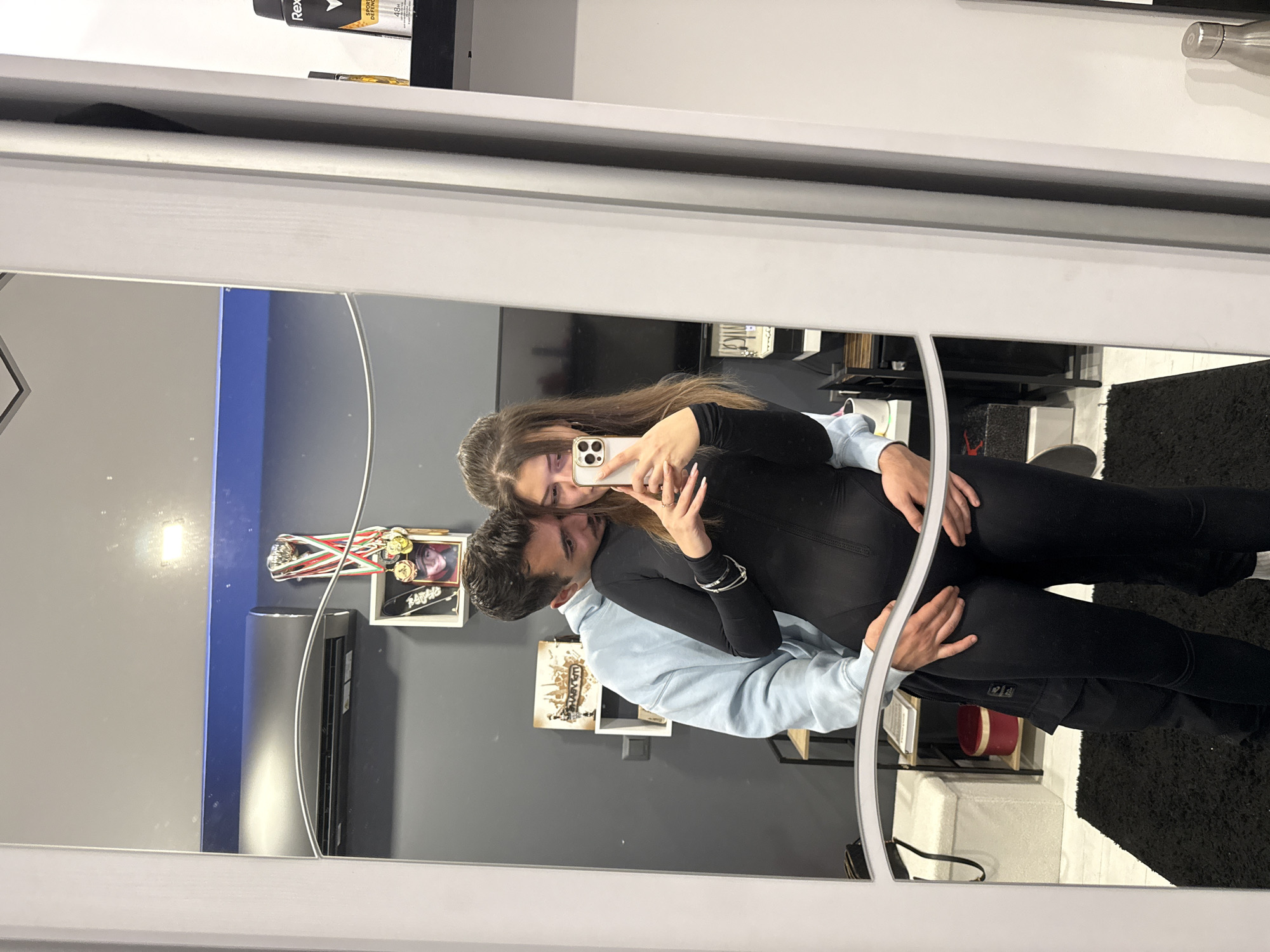A mirrored scene depicts a couple taking a selfie. The woman, holding the phone, has long brown hair, wears a black jumpsuit, and multiple silver bracelets. The man, behind her, wears a light blue hoodie. Their pose suggests intimacy. The background is a room with gray walls, decorations, and shelves. The mirror frame is visible, and part of a shelf with products is seen above the mirror. The black rug appears to be on the right side of the mirrored scene. The location is potentially in Sofia, Bulgaria.