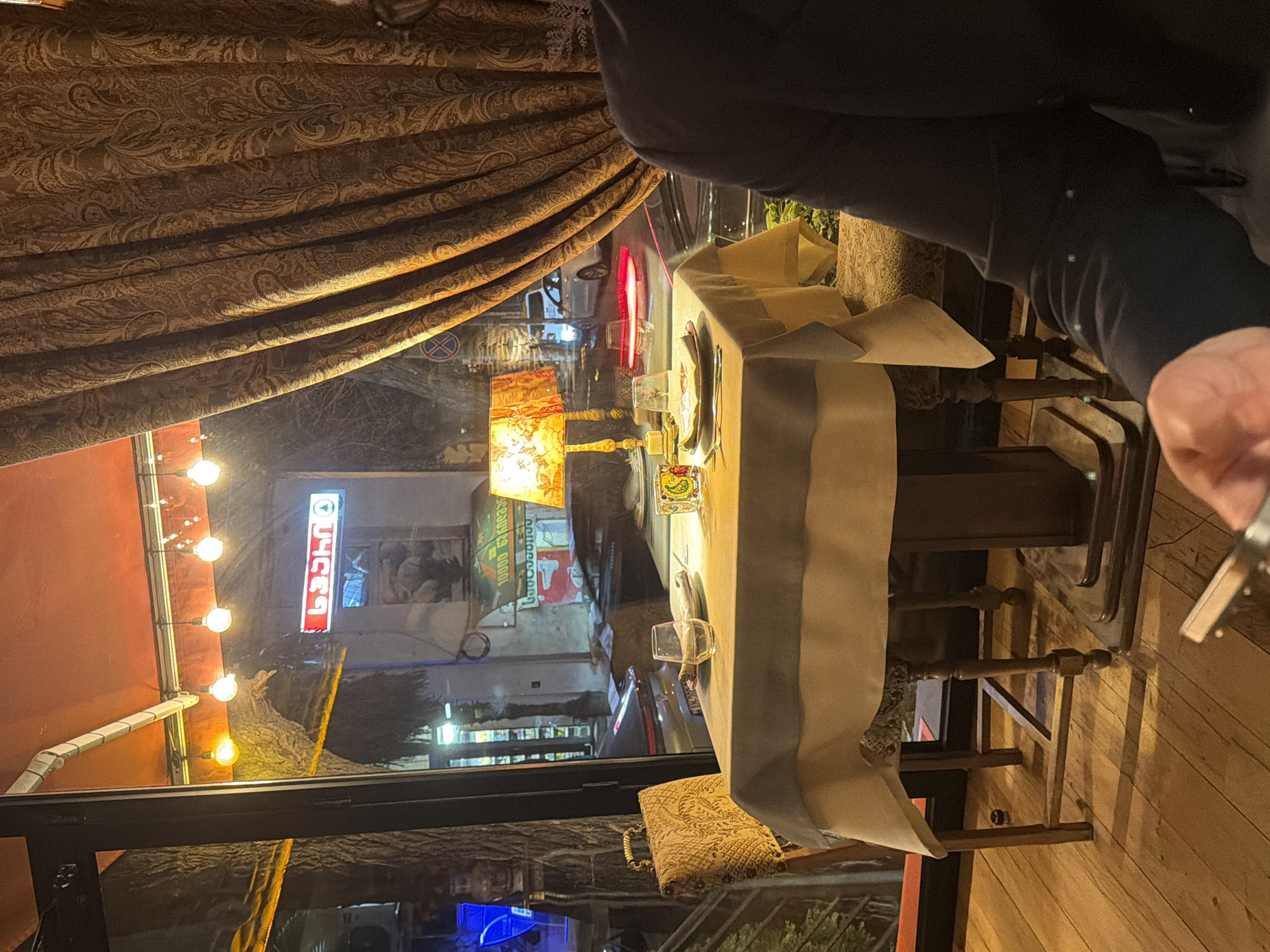 The image depicts an indoor dining scene at a restaurant in Tbilisi, Georgia, at night. The location is inferred from the sign "სპარი" visible through the window, a common convenience store in Georgia. A table is set with a white tablecloth, silverware, and glasses, indicating a meal is in progress. A person in a dark-colored top is partially visible, with a hand holding a lighter. The environment outside the window shows a typical Tbilisi street with storefronts and streetlights. A decorative lamp is lit on the table, and a patterned curtain hangs on the left side of the frame. The setting appears to be a restaurant's interior with an outdoor view.
