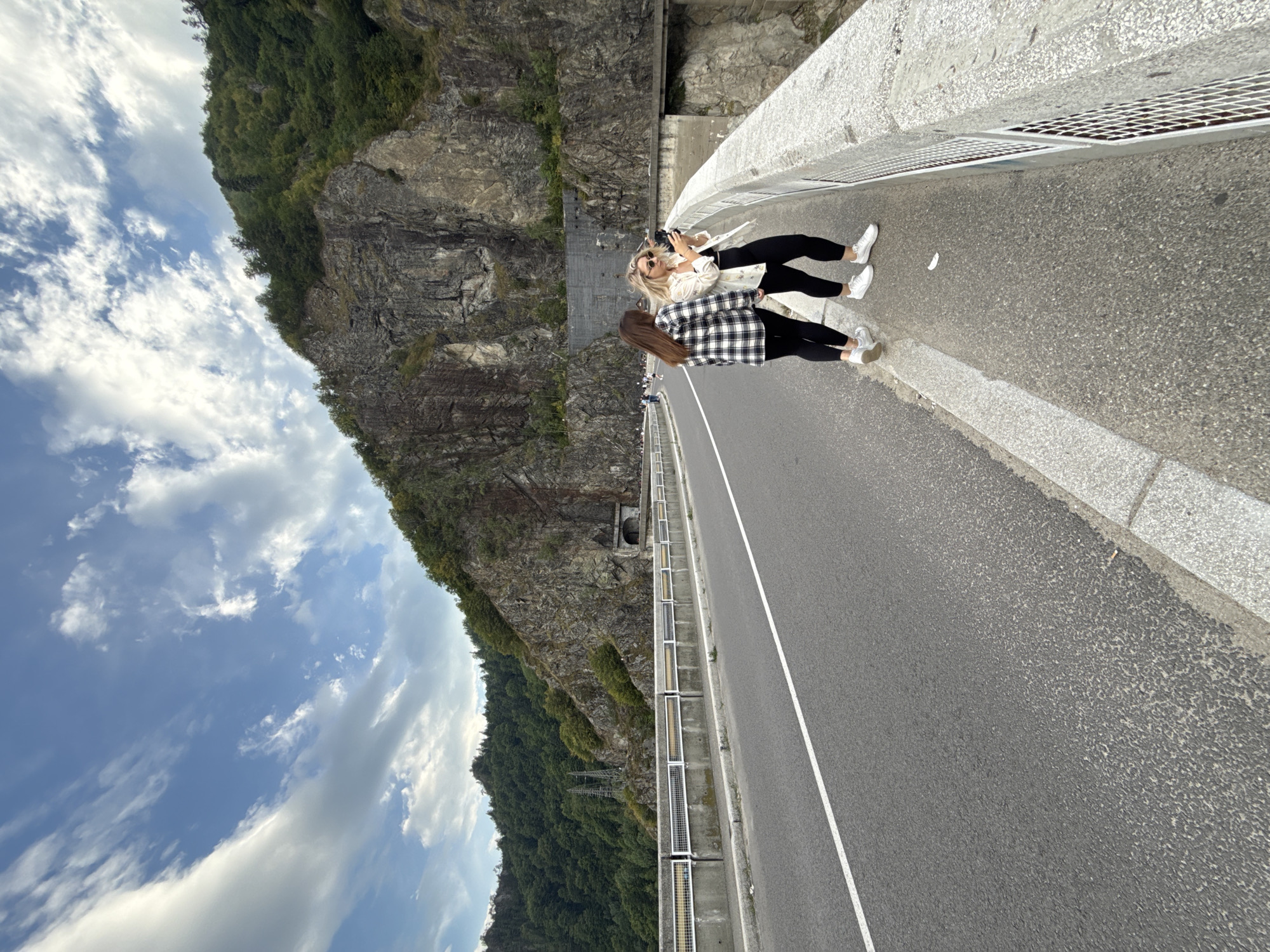 The image captures a view of the Vidraru Dam in Arefu, Romania. Two women are standing on the side of the road atop the dam. The woman on the left wears a black and white checkered shirt and black leggings. The woman on the right, facing the camera, wears a white patterned jacket over a black top and black leggings, and sunglasses. She holds a smartphone in her hands, appearing to take a selfie. The dam itself is constructed from concrete and is bordered by a concrete barrier with metal grate sections. A road runs along the top of the dam, with a white line marking the lane. In the background, a steep, rocky mountainside covered in dense green forest rises dramatically. A tunnel entrance is visible carved into the rock face. The sky above is a bright blue with scattered white clouds.