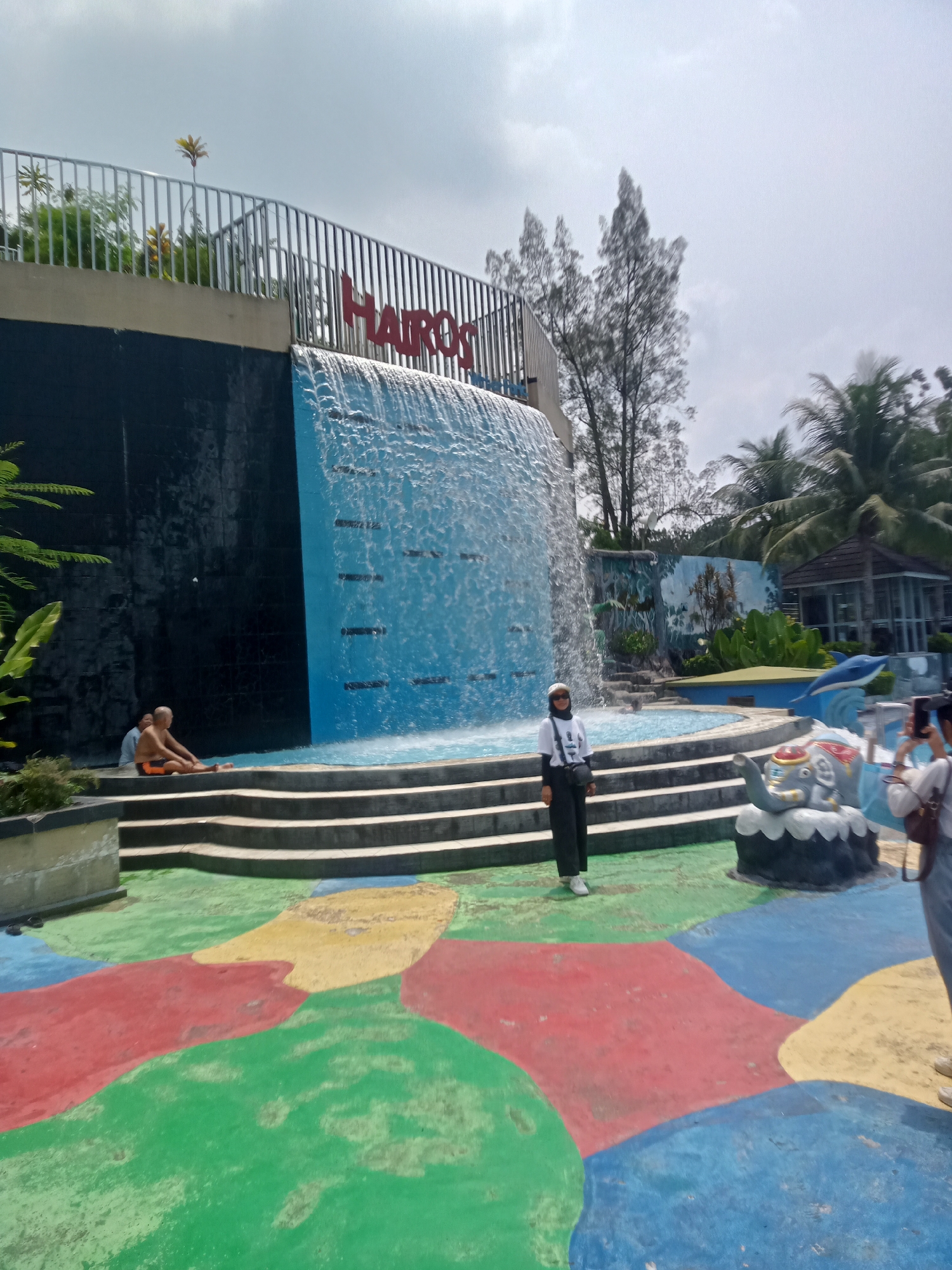 A woman wearing a white hijab, white t-shirt, and black pants stands in front of a man-made waterfall at a water park. The waterfall cascades down a black tiled wall into a blue pool. The woman is looking towards the camera and smiling. To the left of the waterfall, two men are sitting on the steps. The ground in front of the waterfall is painted with colorful abstract shapes in red, green, yellow, and blue. In the background, there are palm trees, a building, and a blue dolphin statue. The sky is cloudy. The scene appears to be in a sunny outdoor location.
