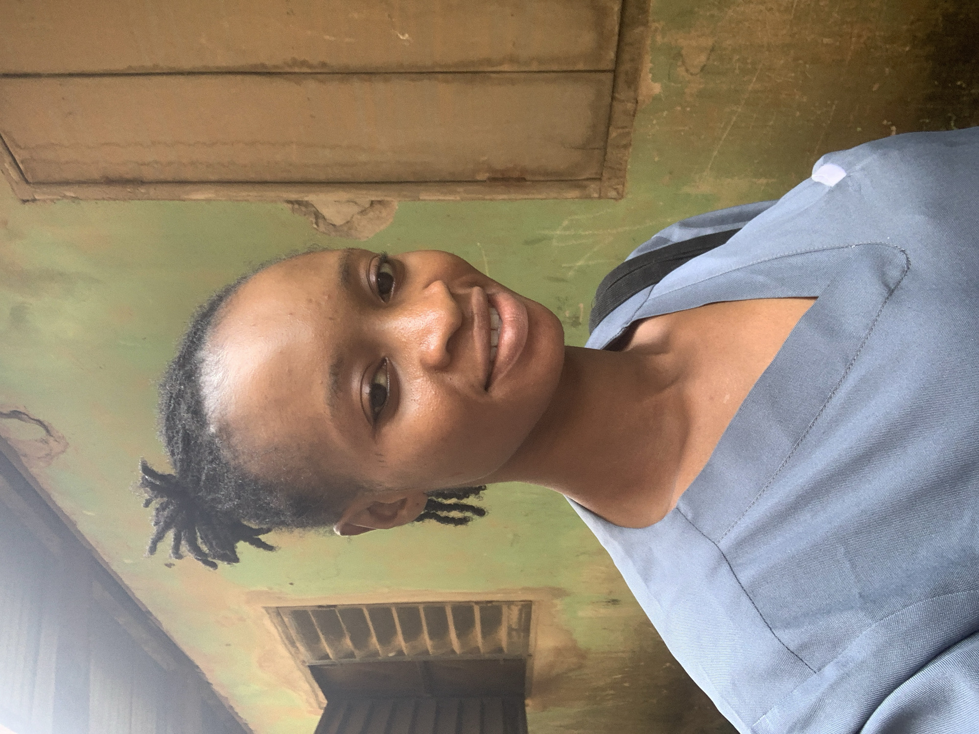 A young Black woman with dreadlocks in a bun smiles at the camera. She is wearing a light blue V-neck scrub top, and a black backpack strap is visible over her left shoulder. She is standing in front of a weathered, green-painted wall with a brown wooden window to her left and a wooden door or shutter to her right. The wall shows signs of age and wear, with chipped paint and dirt. The overall environment appears to be an outdoor setting in a somewhat dilapidated urban area, likely in Shagamu, Nigeria, given the building materials and style.