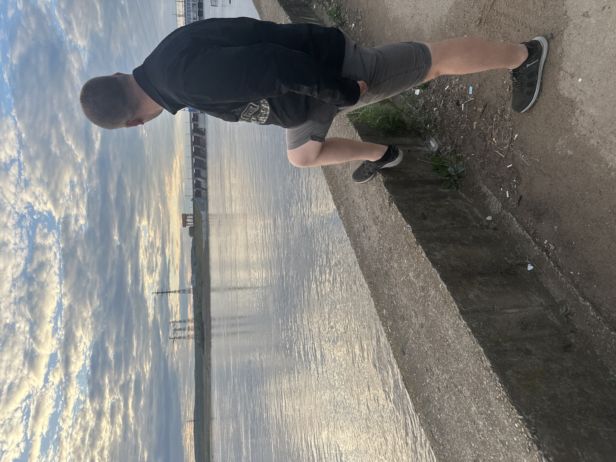 The image shows a person standing at the edge of a body of water, which appears to be a canal or river. The person is wearing a black hoodie and grey shorts, with black athletic shoes. They are looking down towards the water. The scene is set outdoors, with a cloudy sky overhead. The time of day appears to be late afternoon or early evening, as indicated by the soft, diffused light and the golden hues in the clouds.
In the background, there are industrial structures and power lines visible along the opposite bank of the water. The water itself is calm, with ripples reflecting the sky. The ground where the person is standing is concrete, with some debris scattered around.
The overall mood of the image is contemplative and peaceful, with the person seemingly lost in thought while observing the water. The cloudy sky adds a dramatic touch to the scene. There is no visible text in the image.
